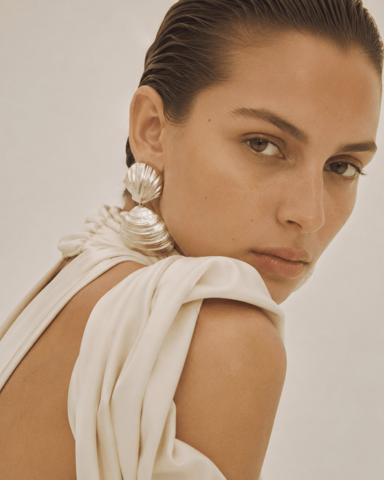 Close-up of a woman with slicked-back hair, wearing large shell-shaped earrings and a cream-colored fabric draped over her shoulder.