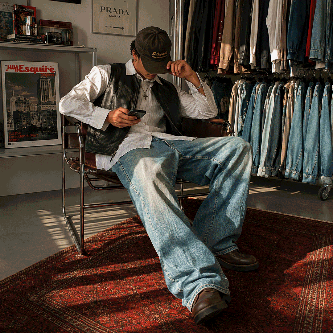 Young man sitting on a vintage chair in a clothing store, looking at his phone, wearing a black cap, leather vest, white shirt, and baggy jeans, surrounded by racks of denim jeans and a magazine stand.