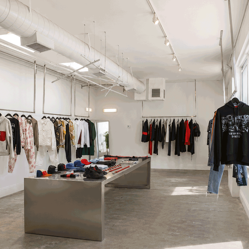 Clothing store with racks of garments, a metal display table with hats, shoes, and accessories, and large windows allowing natural light.