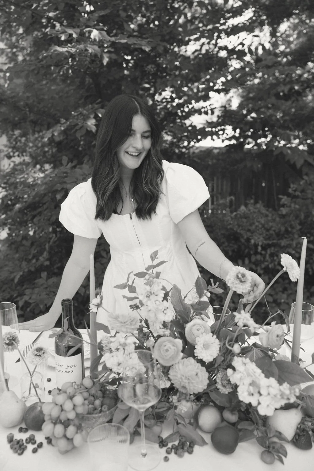 Woman in white dress arranging flowers on a table outdoors.