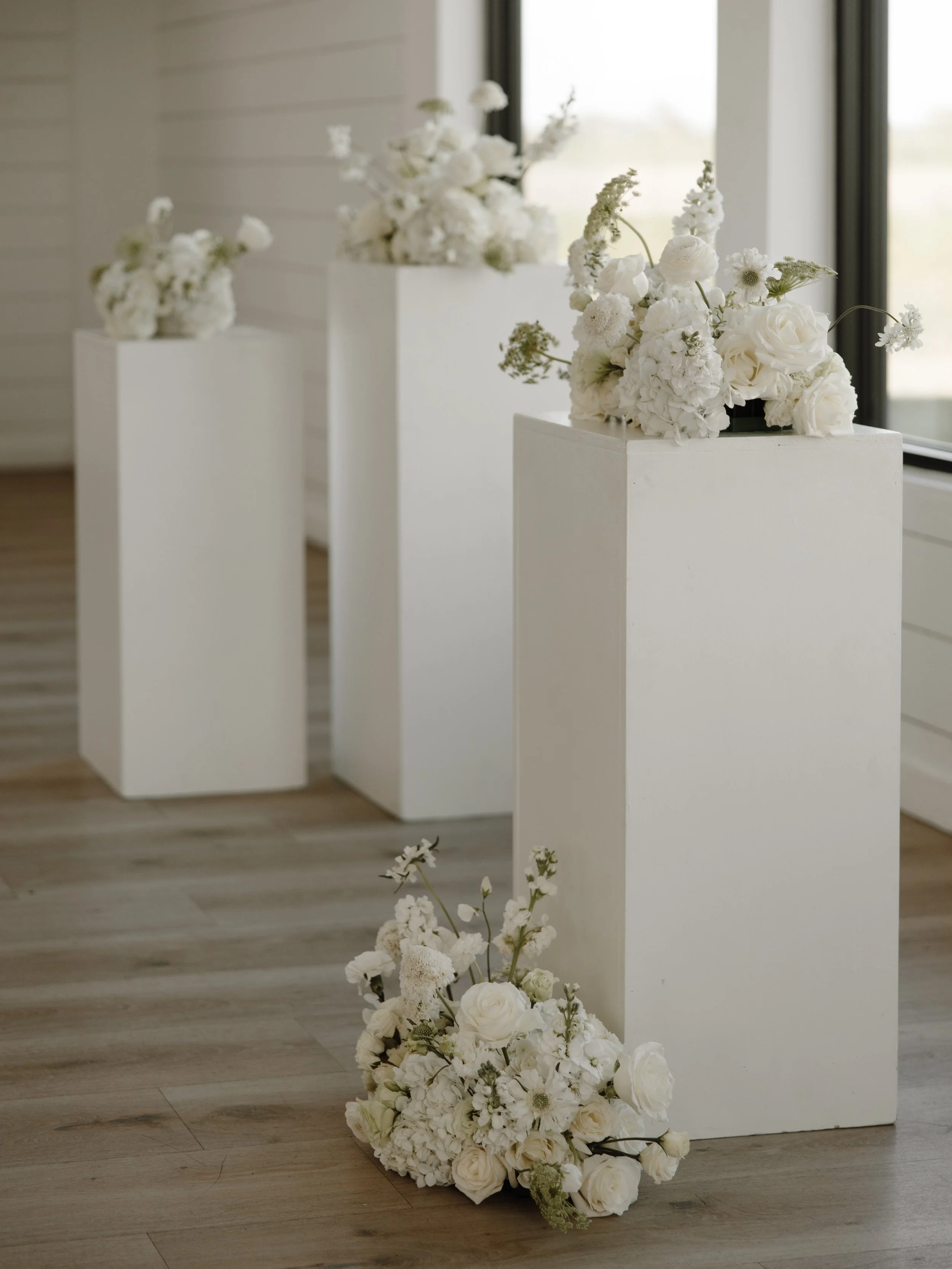 Three white pedestals with arrangements of white flowers displayed in an indoor setting.