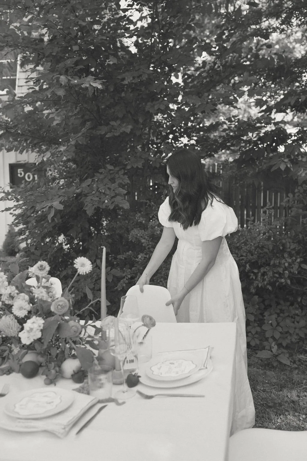 Woman in a white dress setting a table outdoors, surrounded by plants and a floral centerpiece.