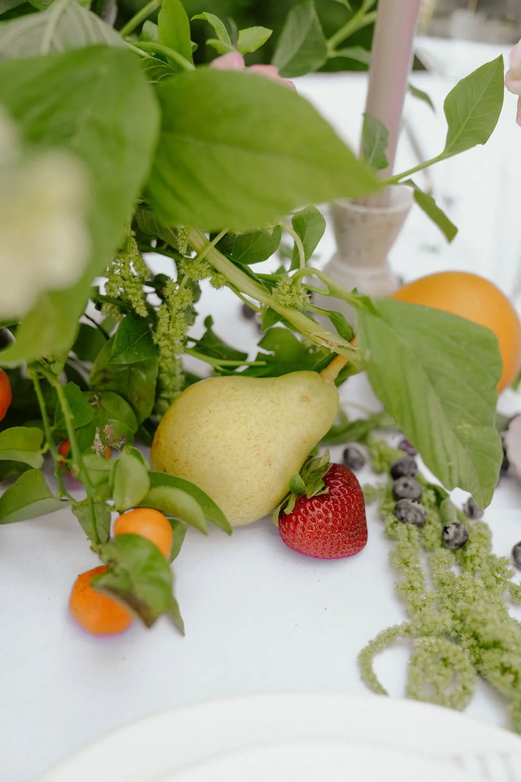 A table setting with a pear, strawberry, kumquats, and blueberries surrounded by green leaves and a candle.