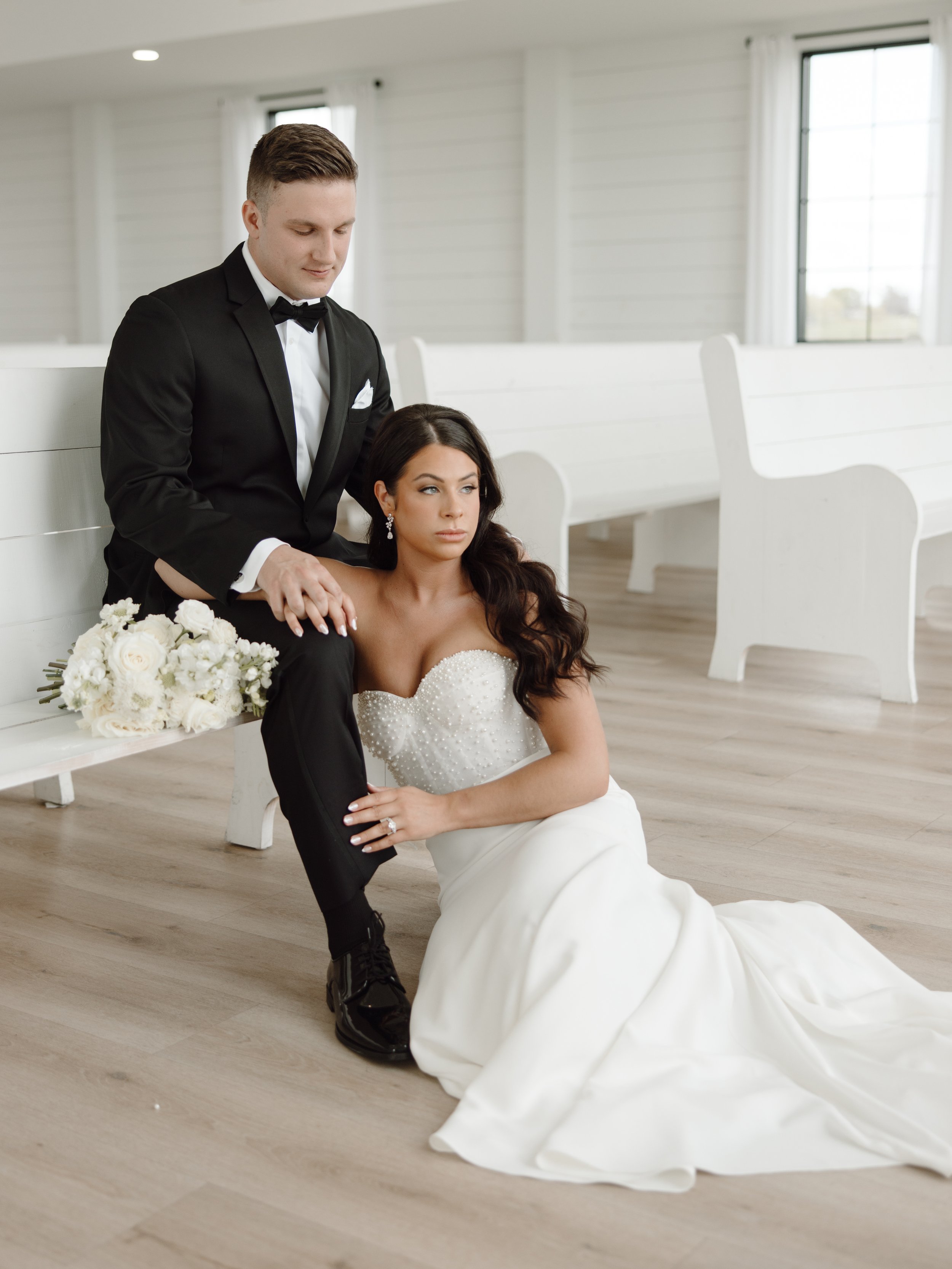 Bride in a white wedding dress sits on the floor next to a groom in a black tuxedo, inside a bright, white chapel.