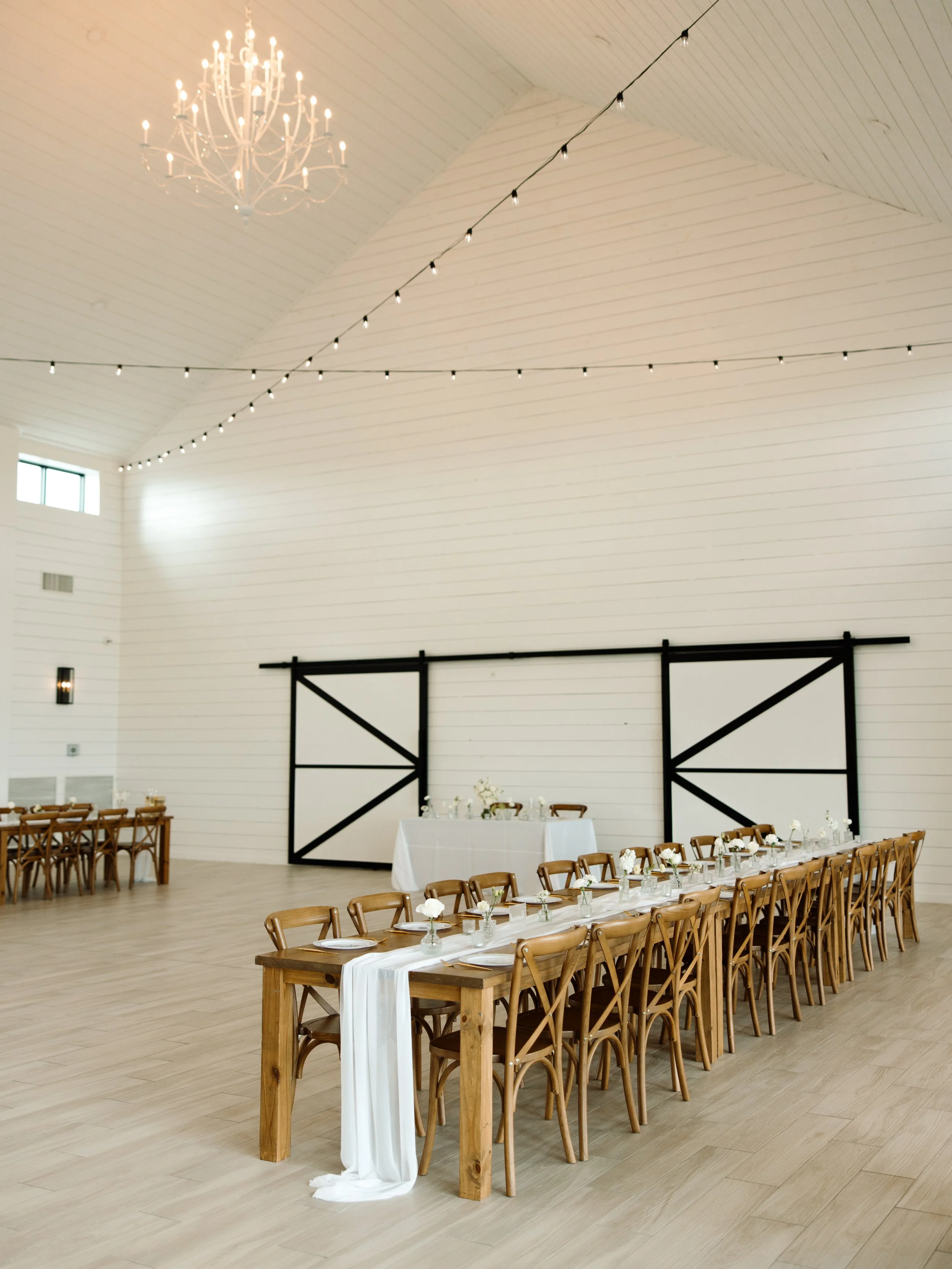 Spacious white barn interior with wooden tables and chairs, decorated for an event with a white table runner. A chandelier and string lights hang from the ceiling, and barn-style doors are visible in the background.