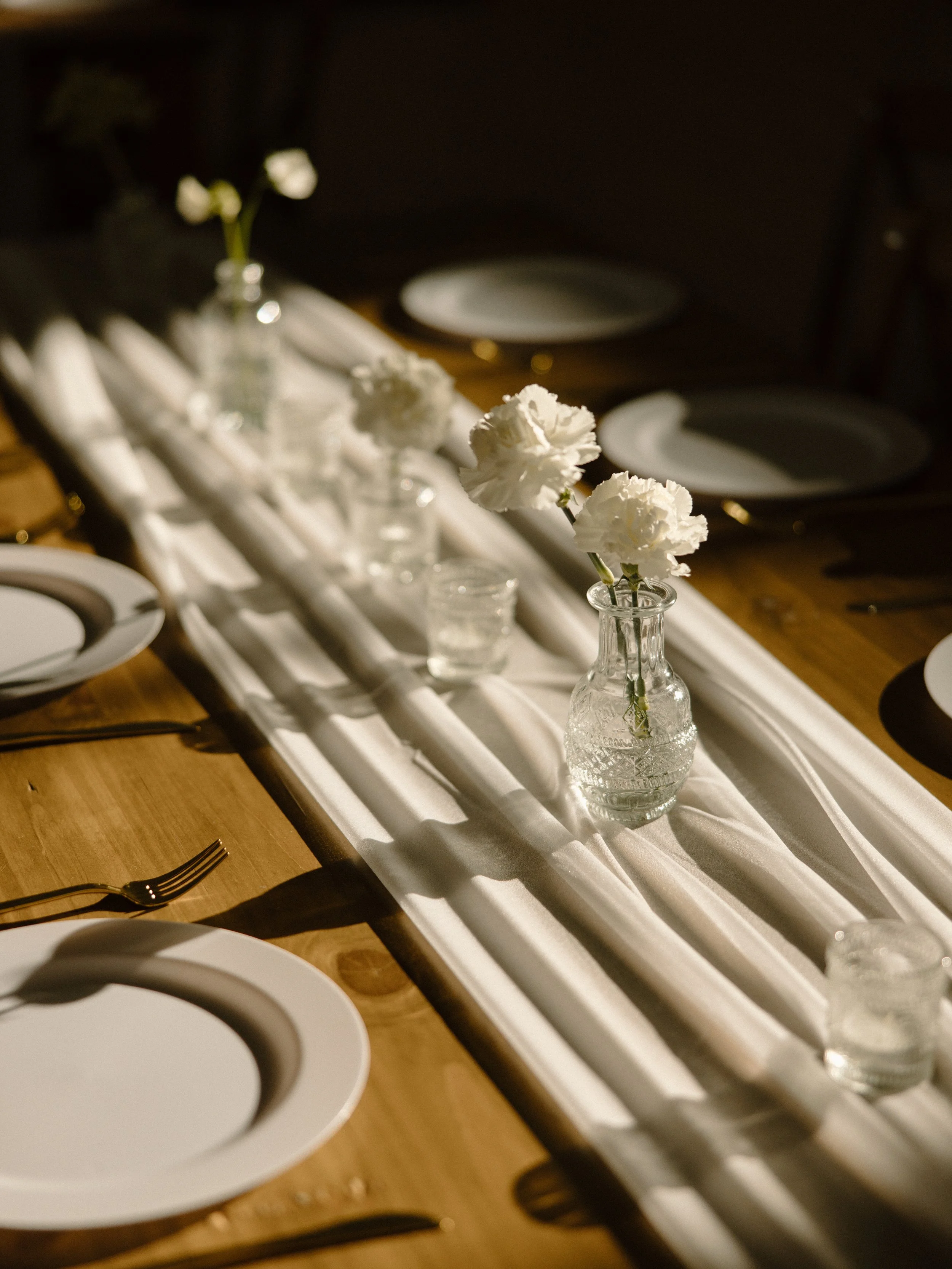 Table setting with white plates, gold cutlery, white flowers in glass vases, and a white table runner.
