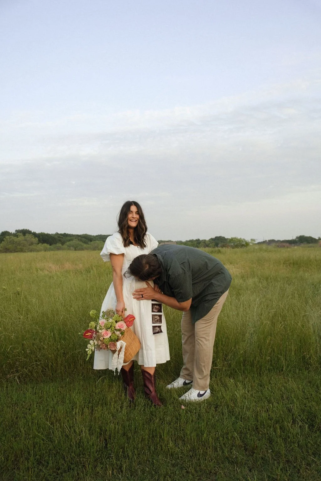 Couple in a field with woman holding sonogram photos, man kissing her belly.