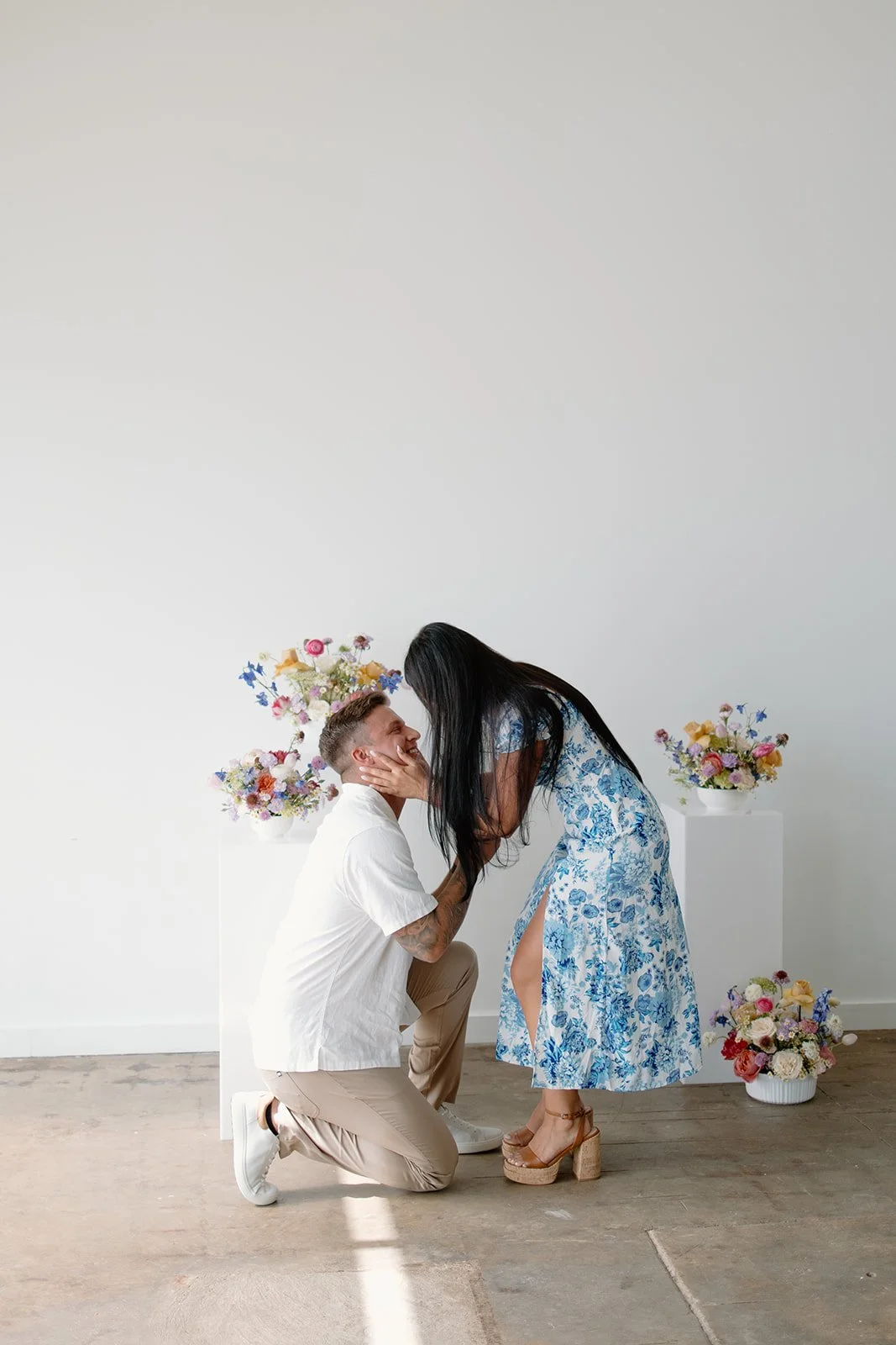 A woman in a blue floral dress kisses a kneeling man in a white shirt and khaki pants. They are surrounded by floral arrangements on white pedestals.