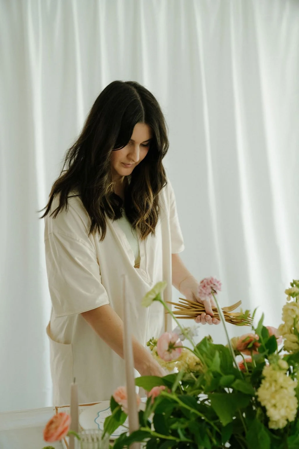 Person arranging flowers at a table with candles, green leaves, and pink blossoms, in front of a white curtain.