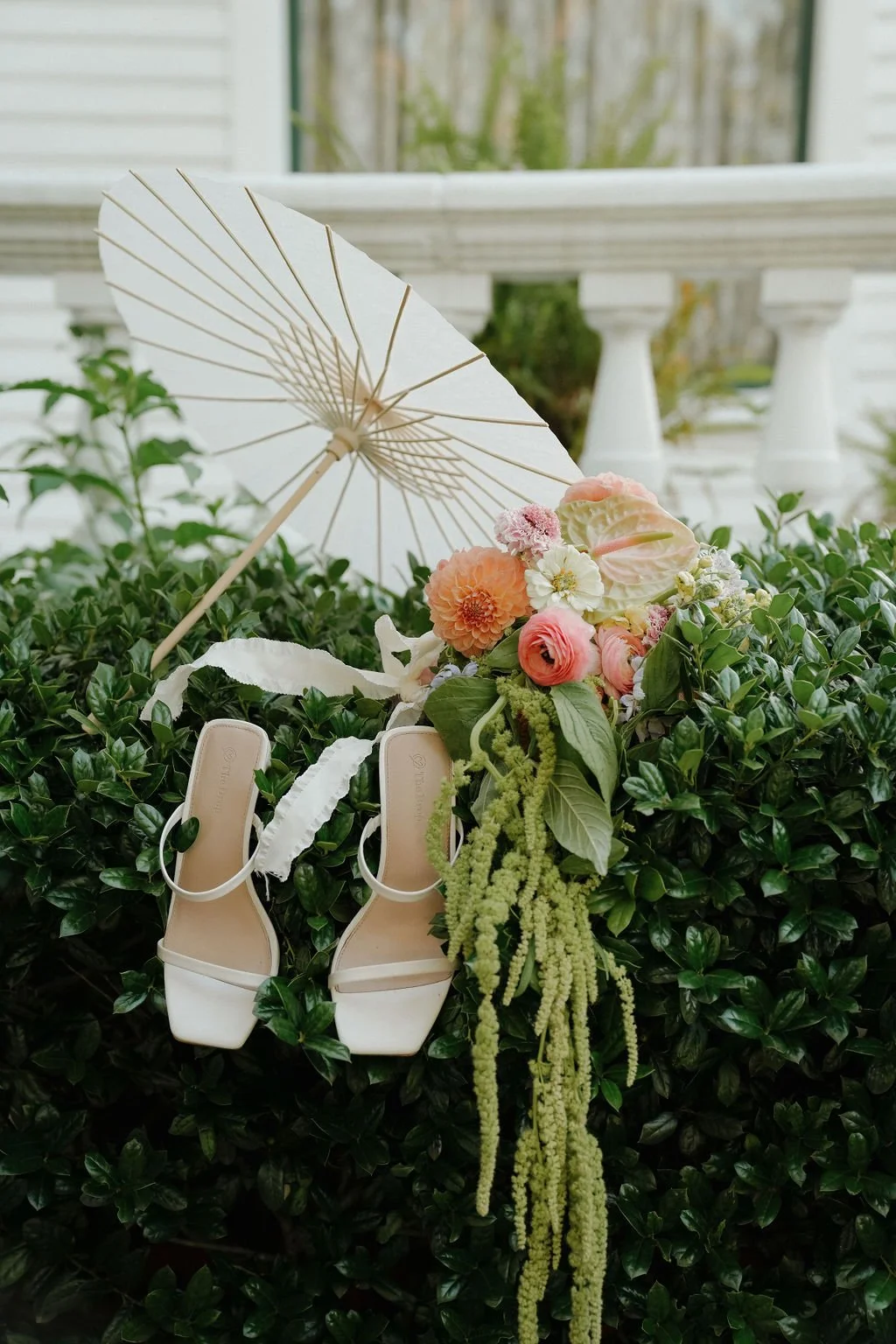 White high-heeled sandals, a paper parasol, and a colorful floral arrangement on green foliage.