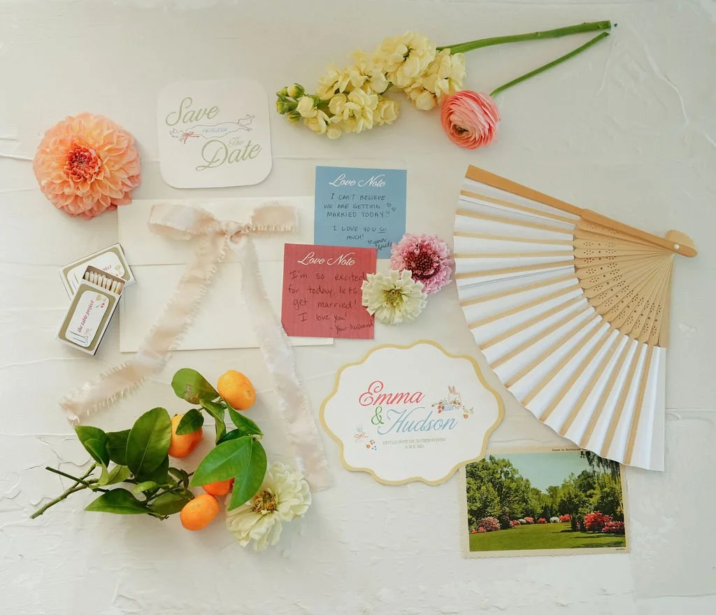 Flat lay of wedding decoration items including a "Save the Date" card, note cards, flowers, ribbon, fan, matchbox, sprig with fruits, and a name card with floral design.