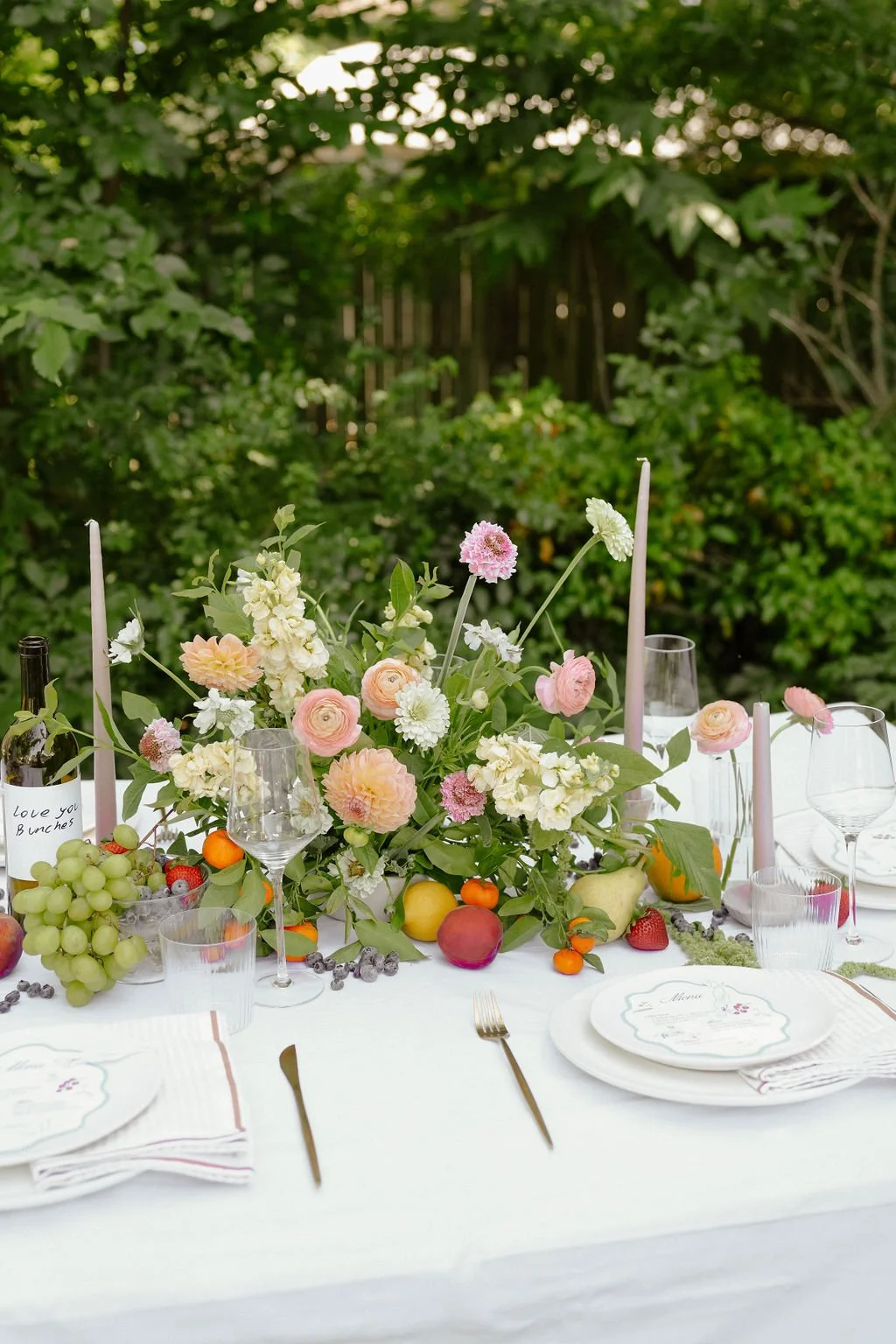 Outdoor dining table with floral centerpiece, fruit, wine bottle, and glassware.