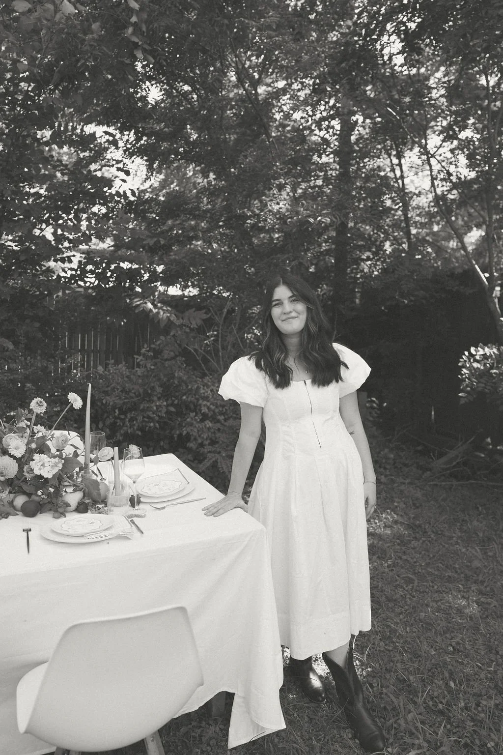 Woman in white dress standing next to a table set with plates and flowers in a garden.