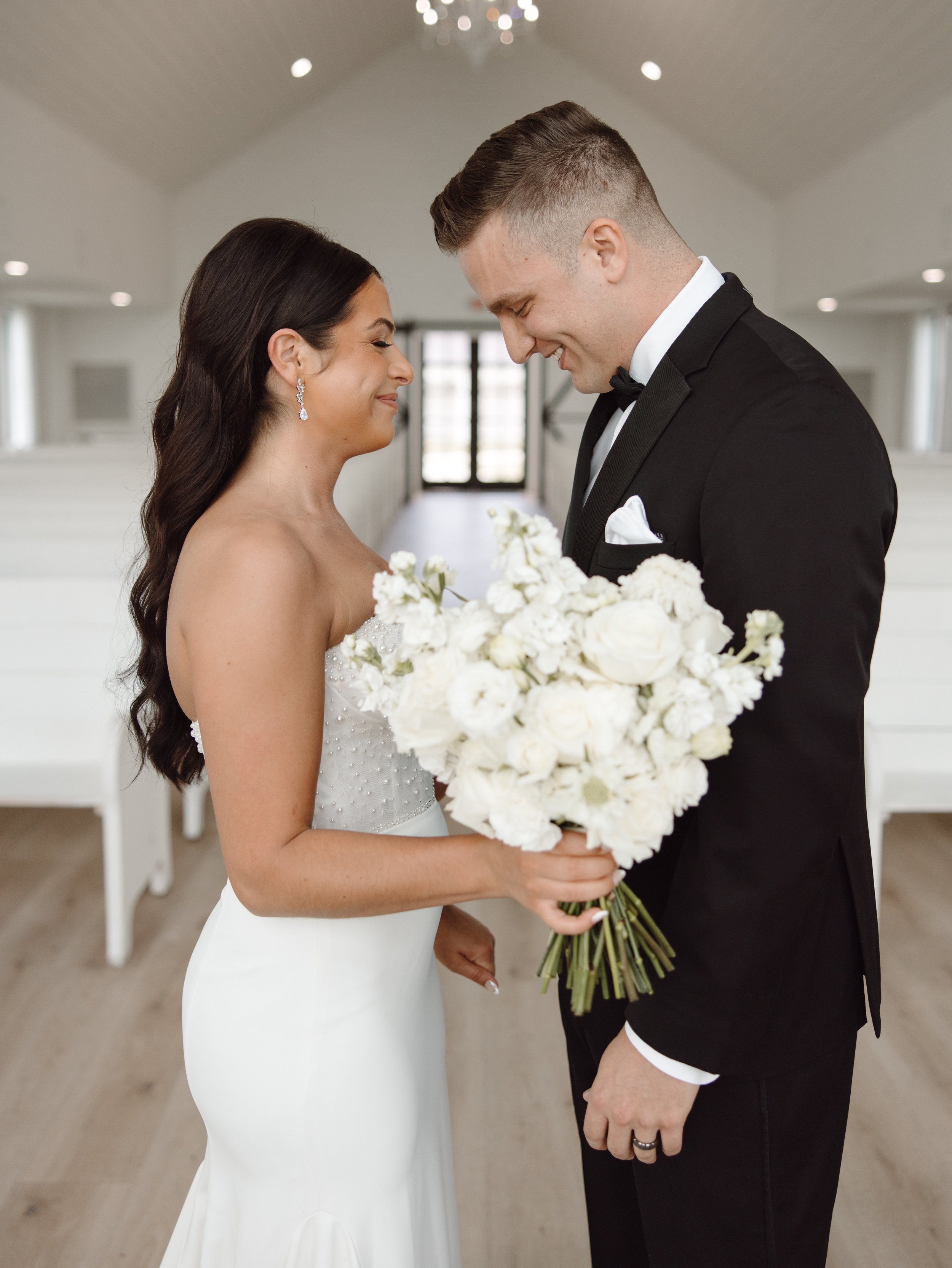 Bride and groom smiling at each other inside a chapel, bride holding white bouquet.