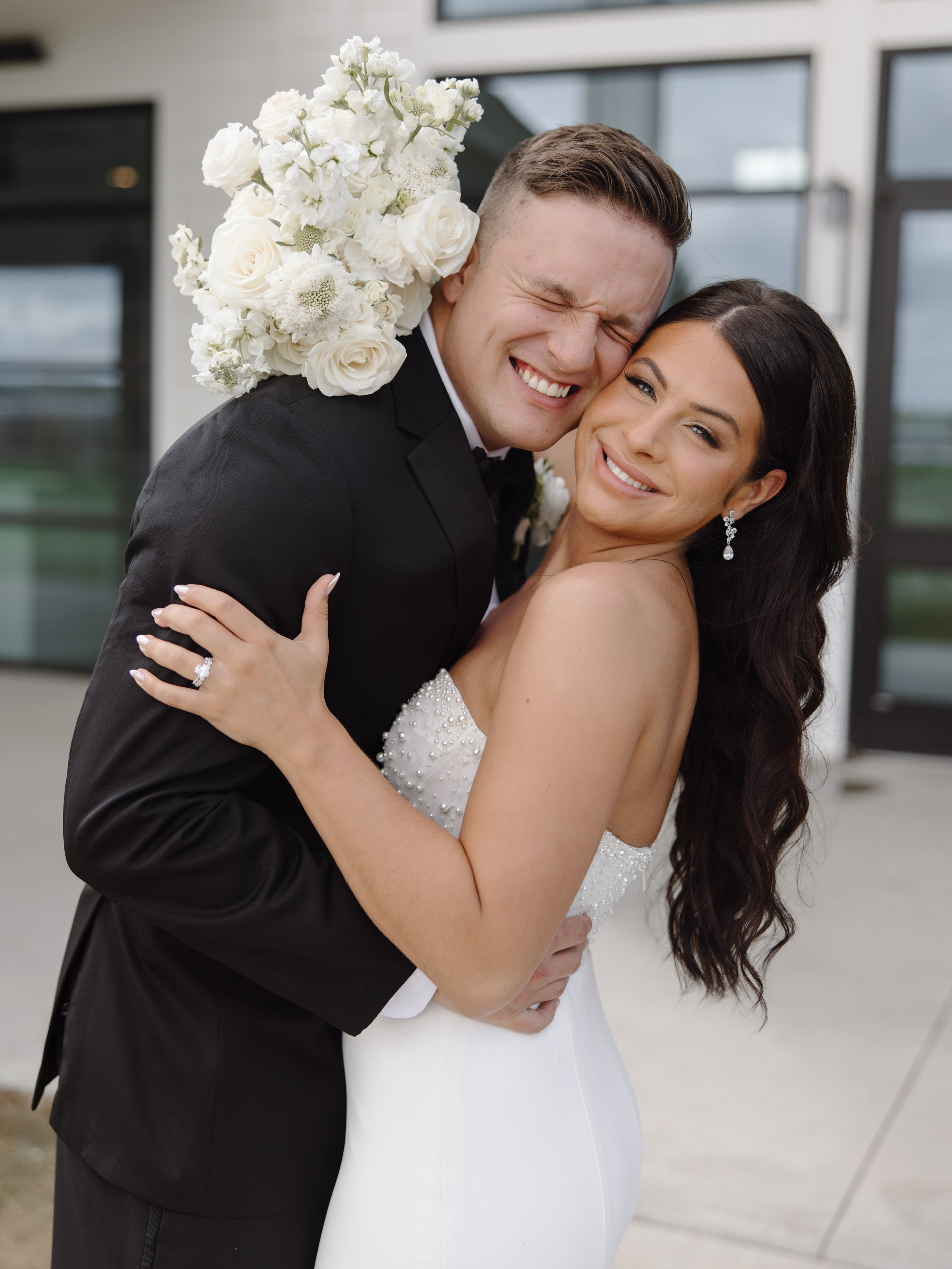 Smiling bride and groom embracing, groom holding bouquet of white flowers.