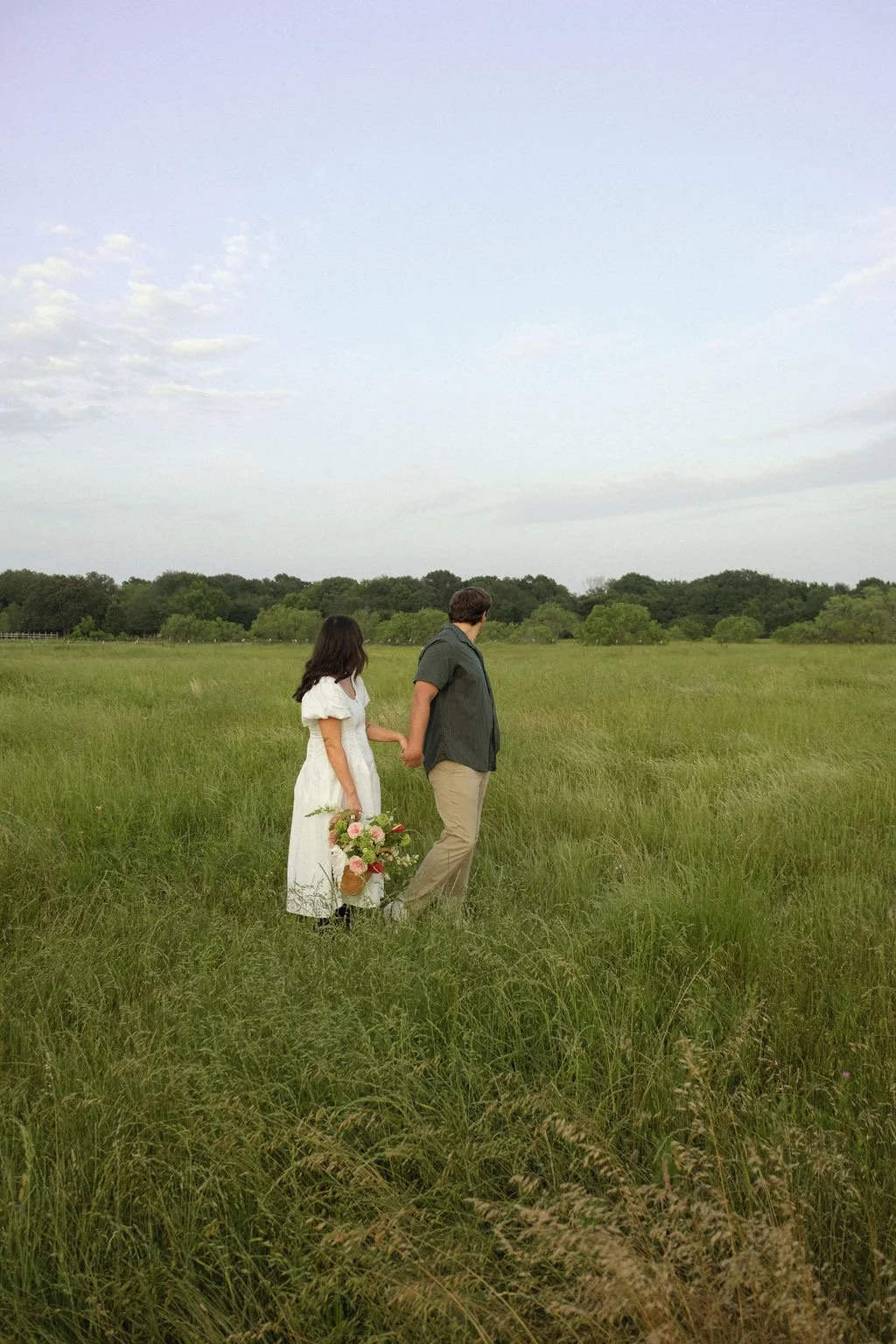 A couple walking through a grassy field holding hands, with the woman holding a bouquet of flowers.