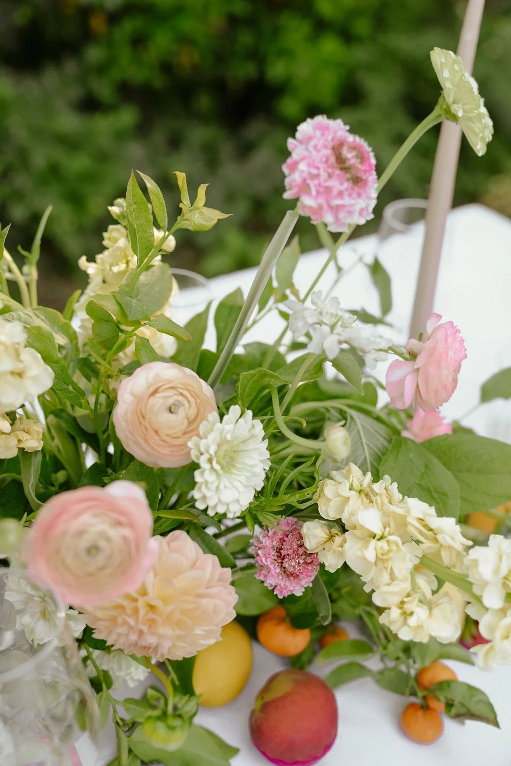 Close-up of a floral arrangement with pink and white flowers, green leaves, and assorted fruits on a table.