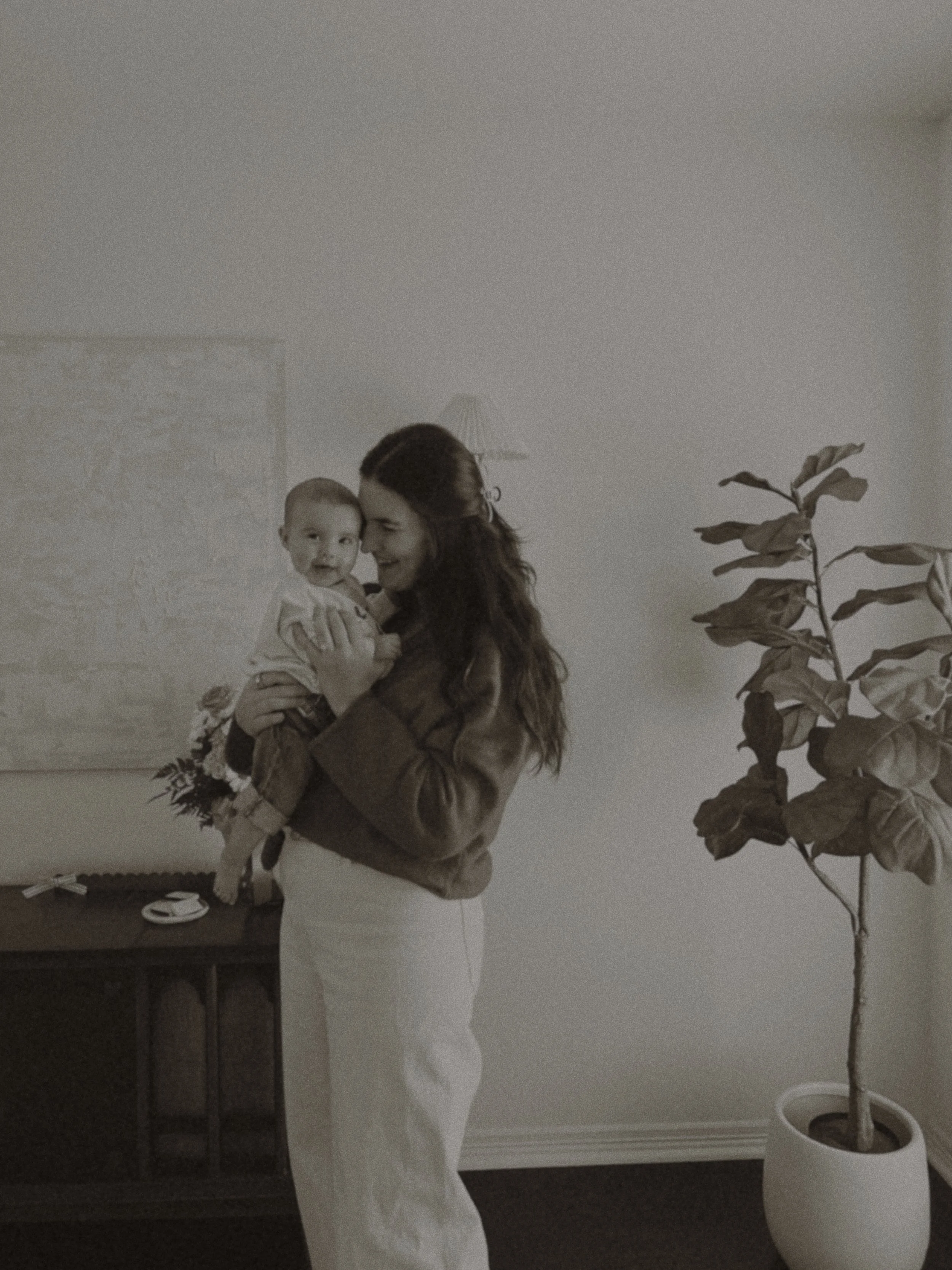 A woman holding a baby in a warmly lit room next to a potted plant and a wooden cabinet.