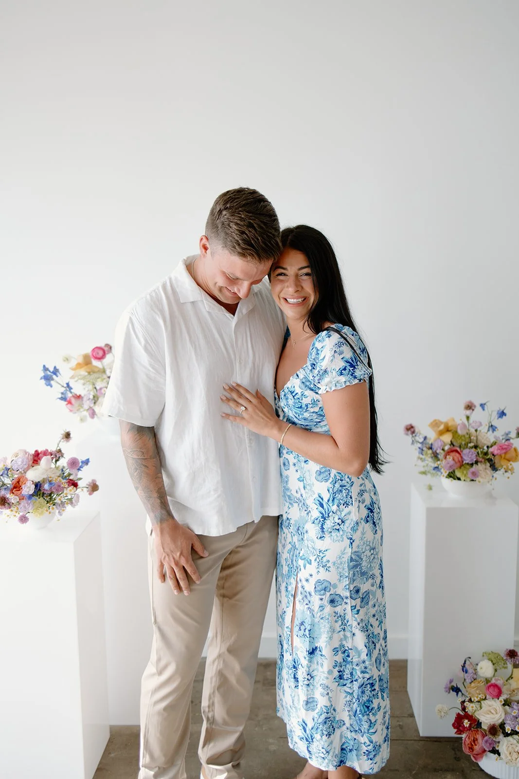A couple in a light room with flower arrangements, the woman wearing a blue floral dress and the man in a white shirt and beige pants.
