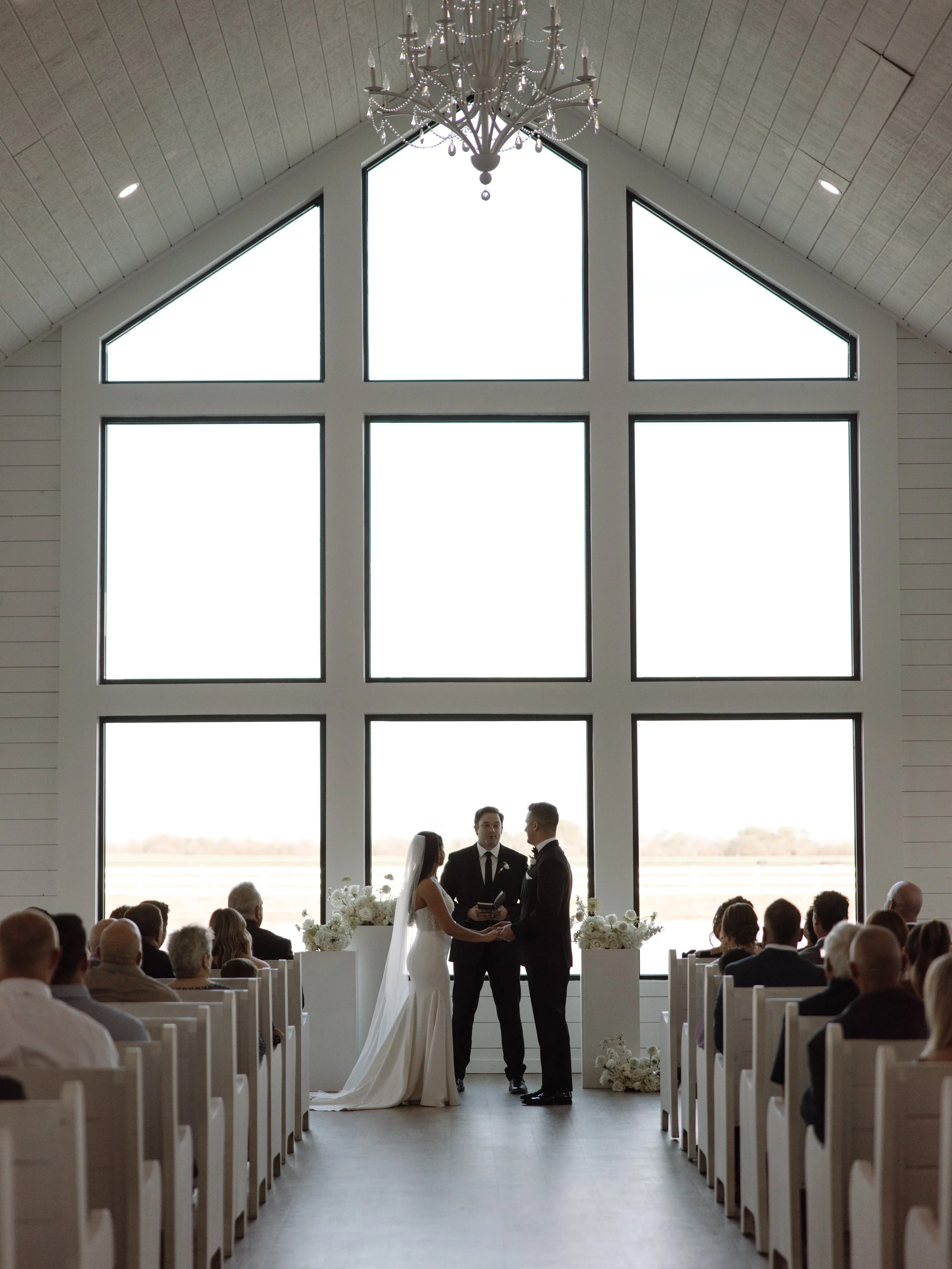 A wedding ceremony in a chapel with large windows, a bride and groom stand before an officiant, witnessed by seated guests. A chandelier hangs from the ceiling.