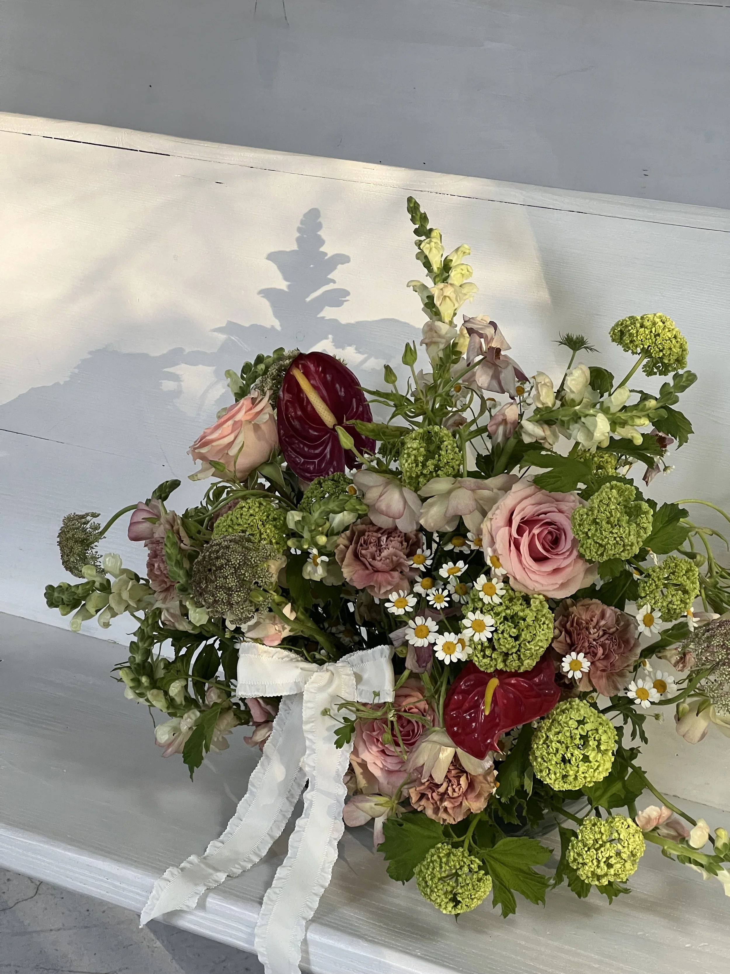 Bouquet of mixed flowers with pink roses, green hydrangeas, red anthuriums, white daisies, and a white ribbon on a light surface.