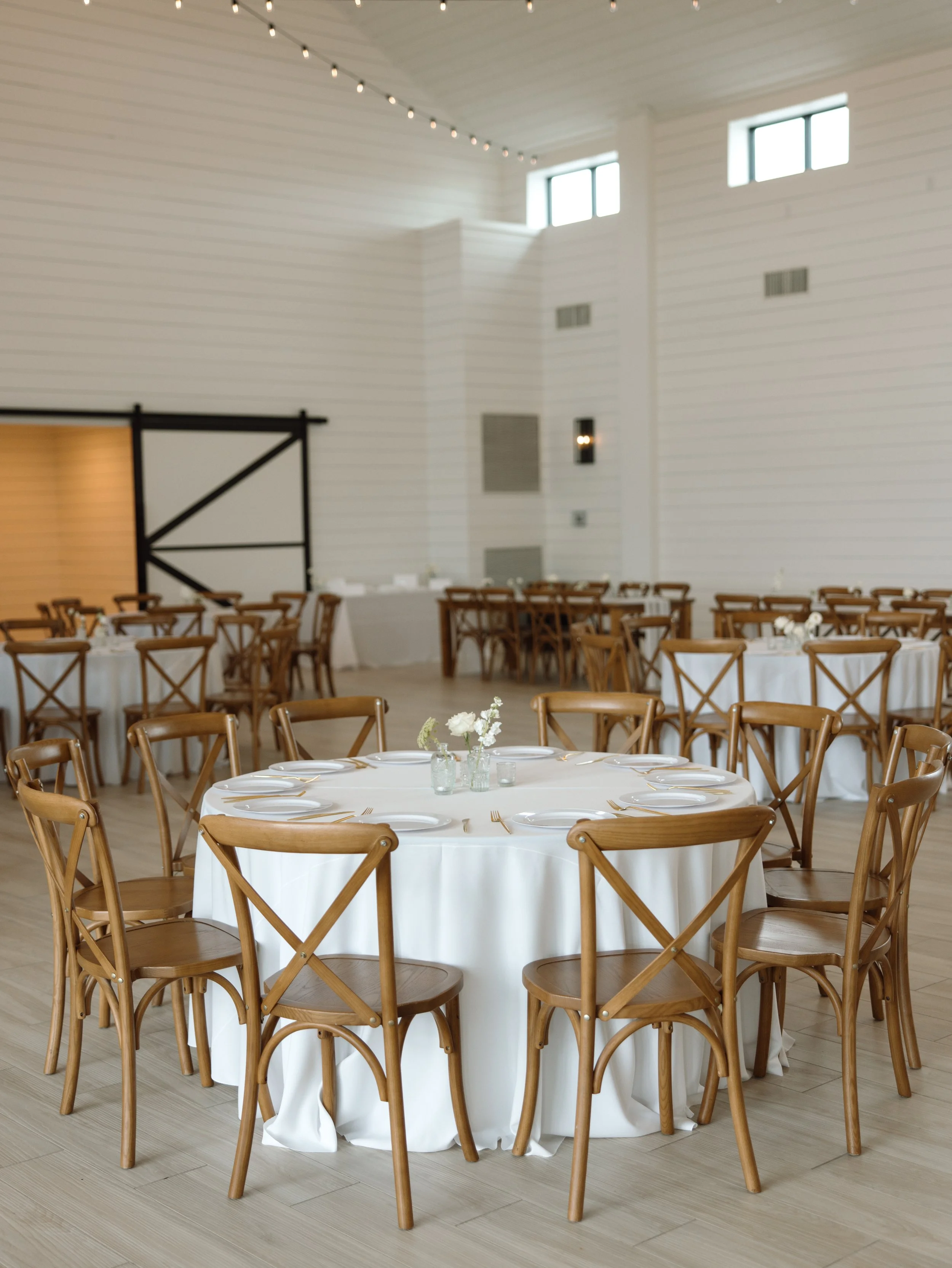 Elegant event space with round tables covered in white tablecloths, wooden chairs, and minimal floral centerpieces, in a room with white walls and string lights overhead.