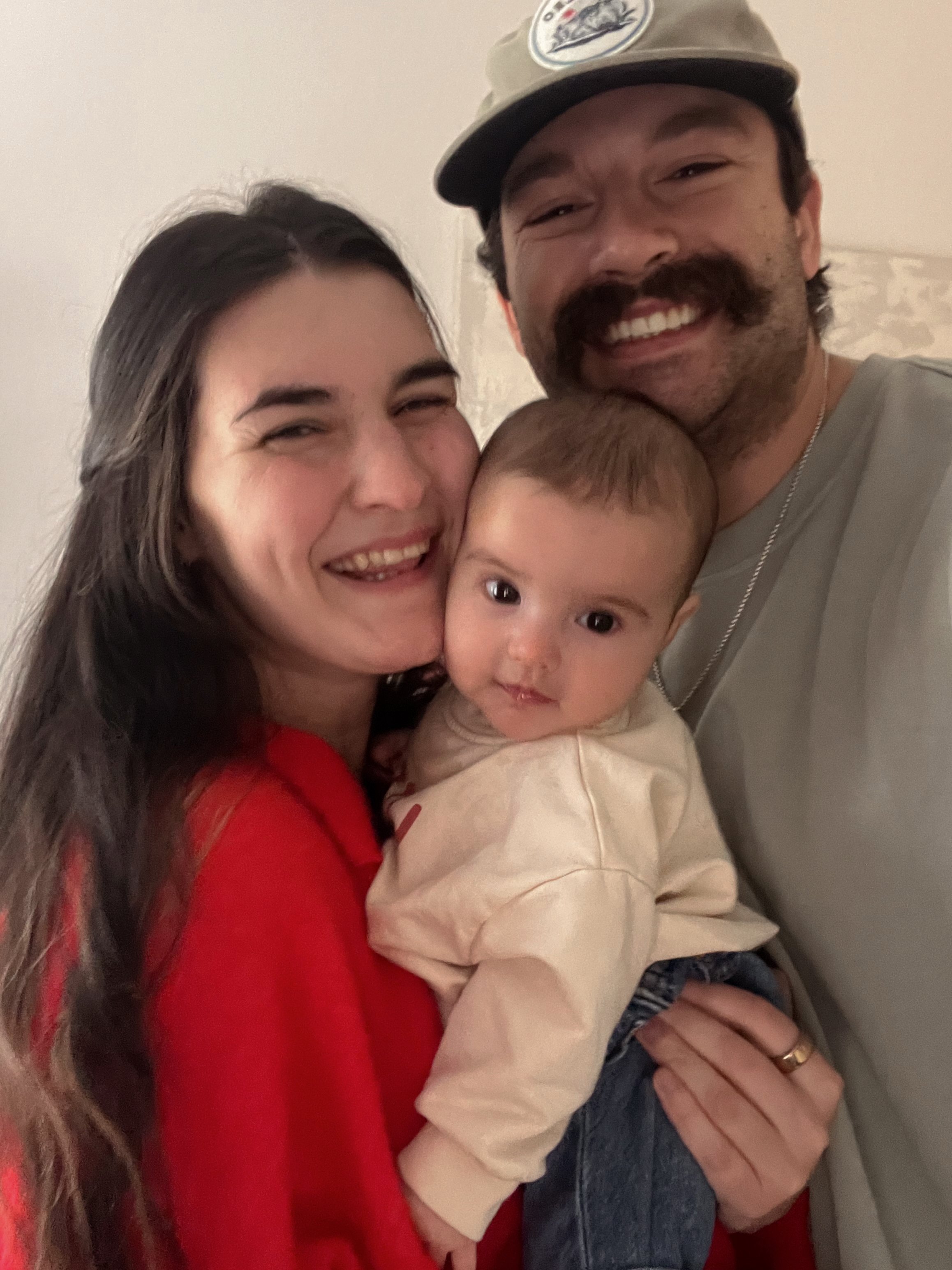 Smiling couple holding a baby, with the man wearing a cap and the woman in a red sweater.