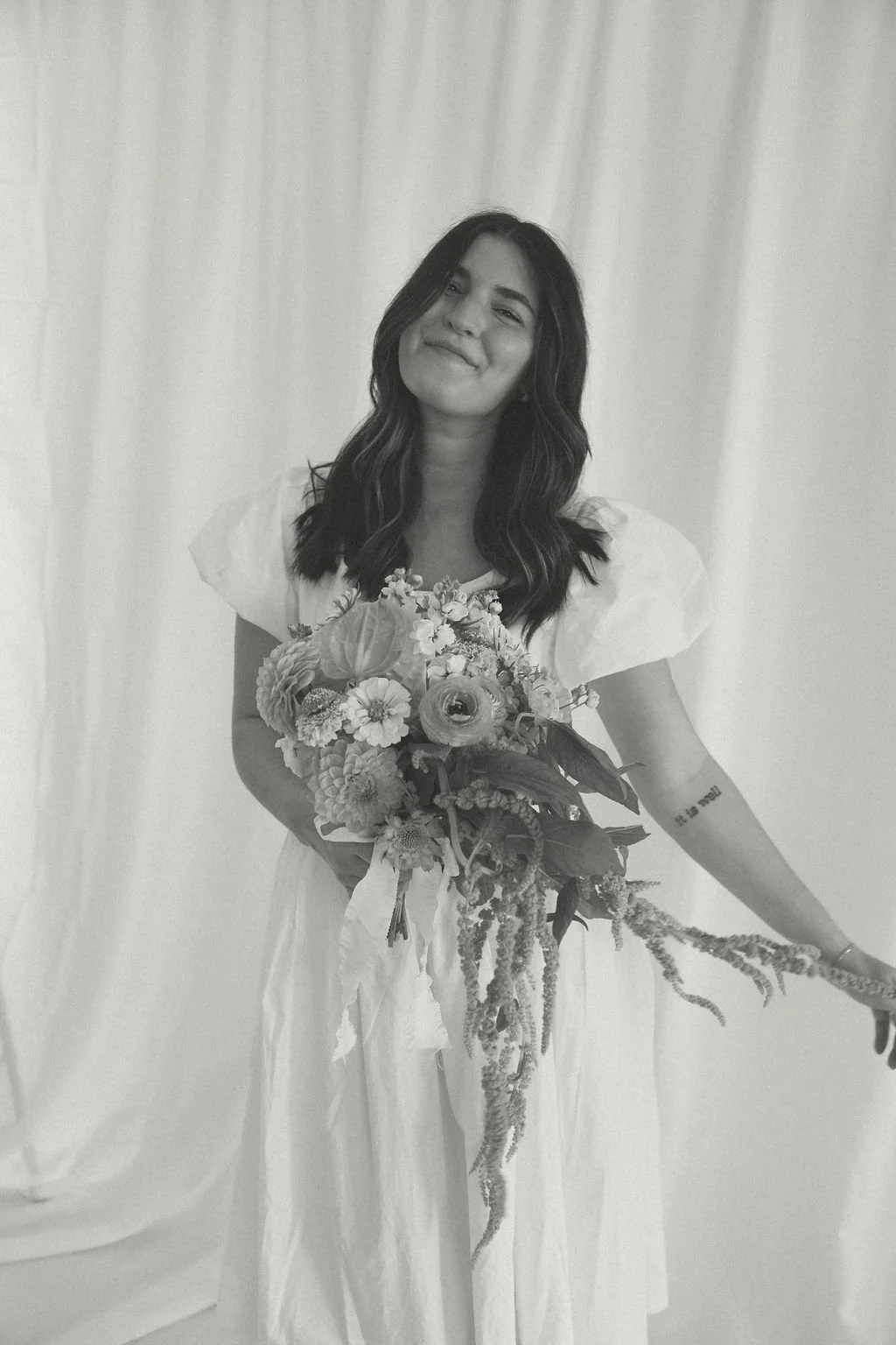 Woman in a white dress holding a bouquet of flowers, standing against a plain backdrop.