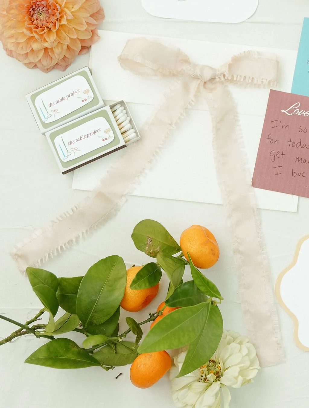 Floral arrangement with oranges, a pink bow, and matchboxes labeled "the table project" on a textured surface.