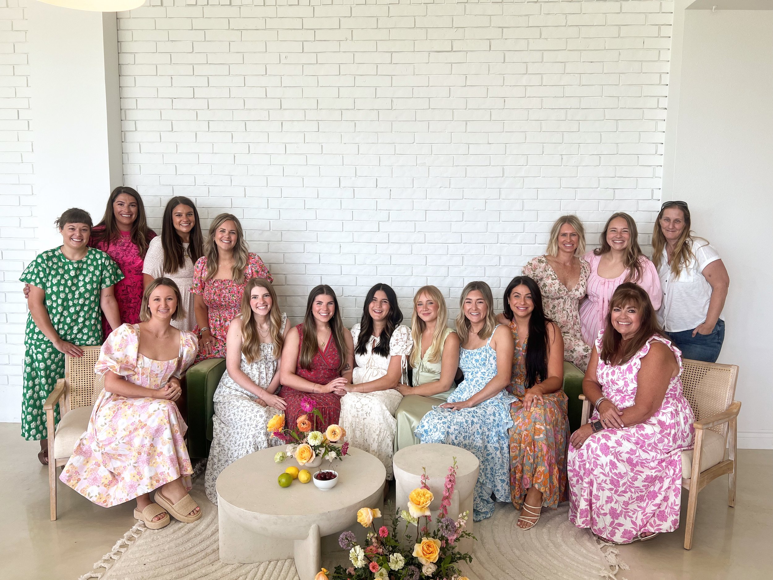 A group of women smiling and posing indoors in front of a white brick wall, wearing colorful dresses, with a floral arrangement and small table in the foreground.