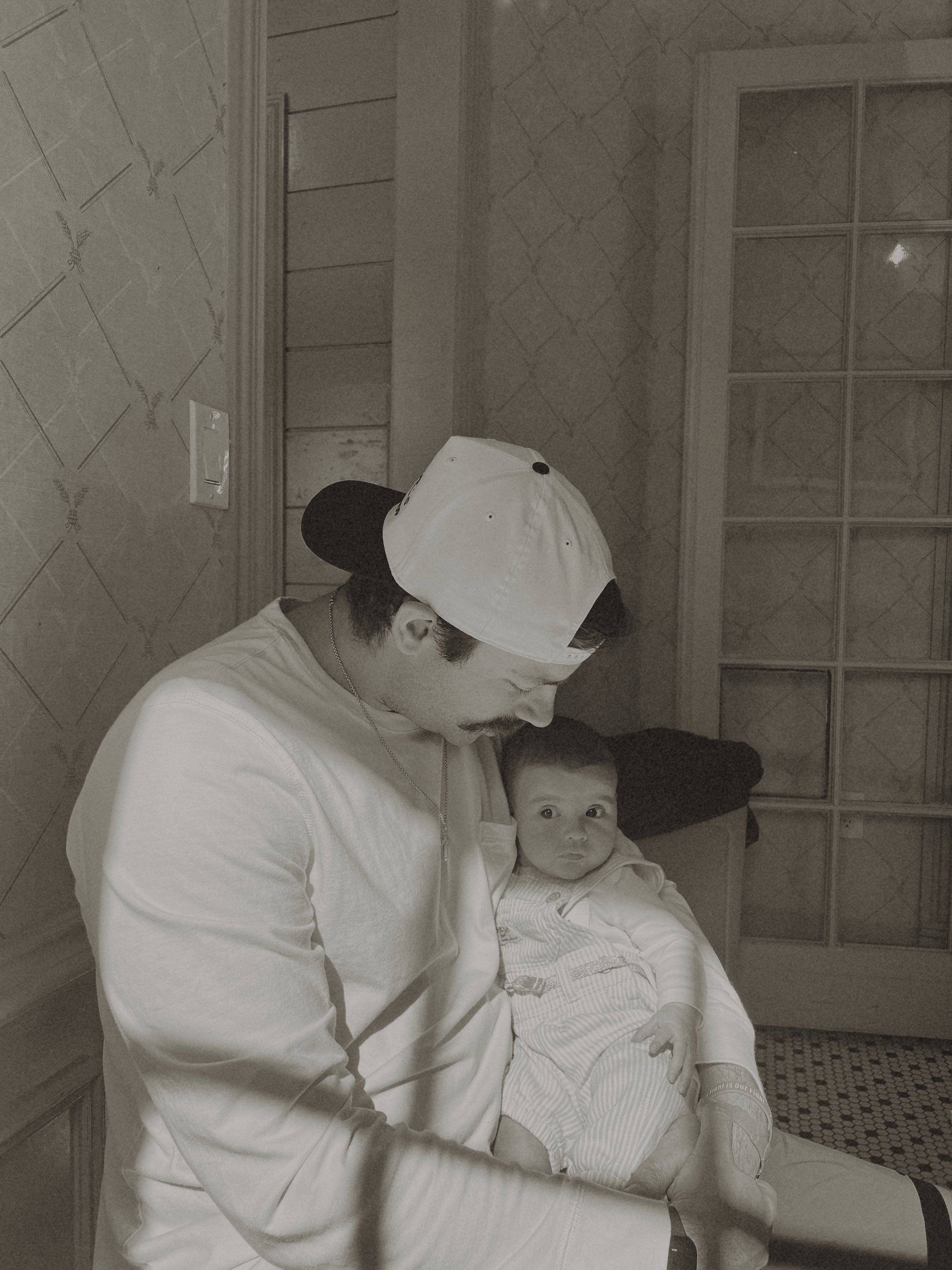 A man wearing a backward baseball cap holding a baby.