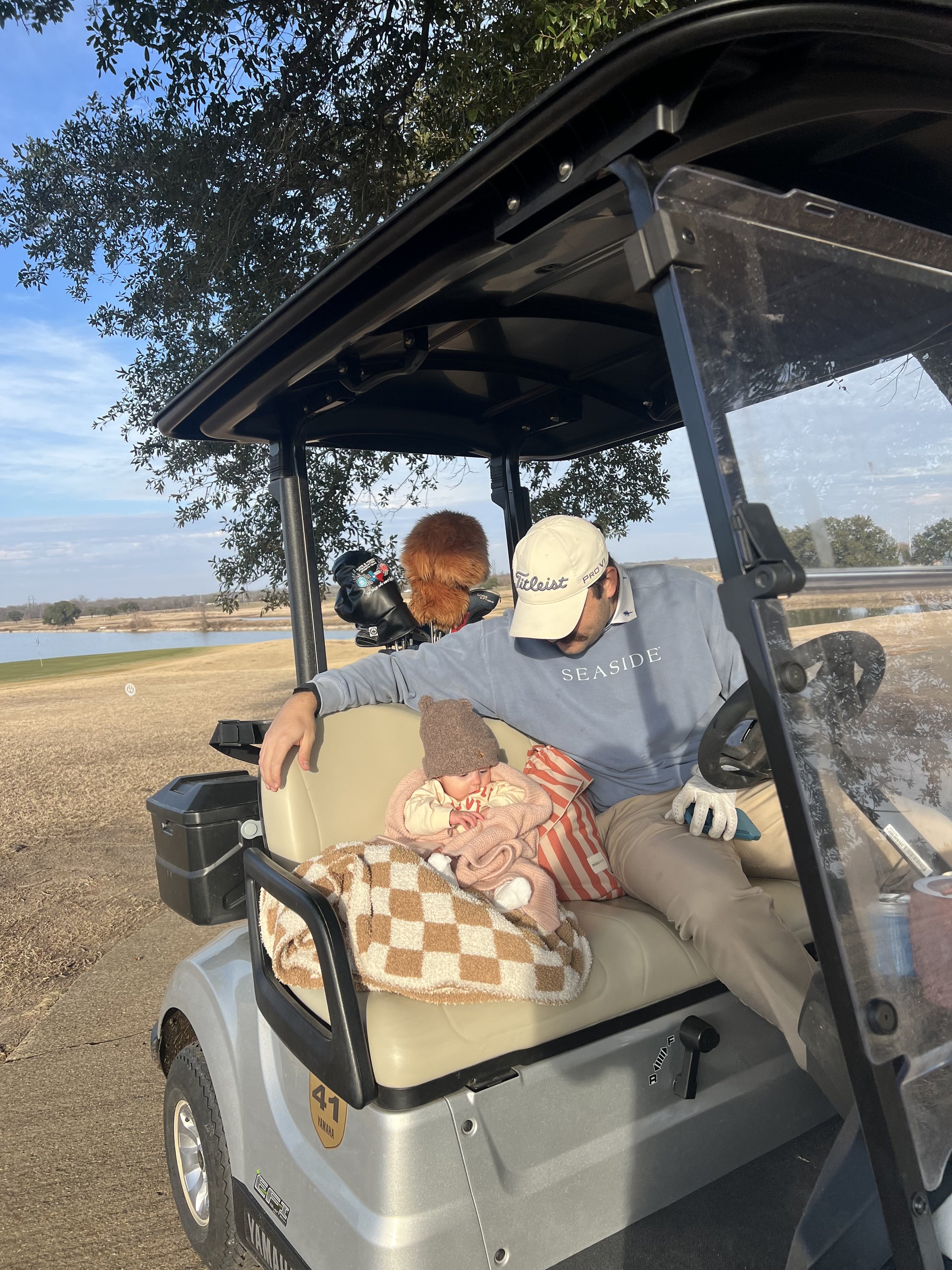 Person sitting in a golf cart with a baby wrapped in a blanket, alongside a golf bag with clubs. Scenic outdoor setting with trees and a lake in the background.