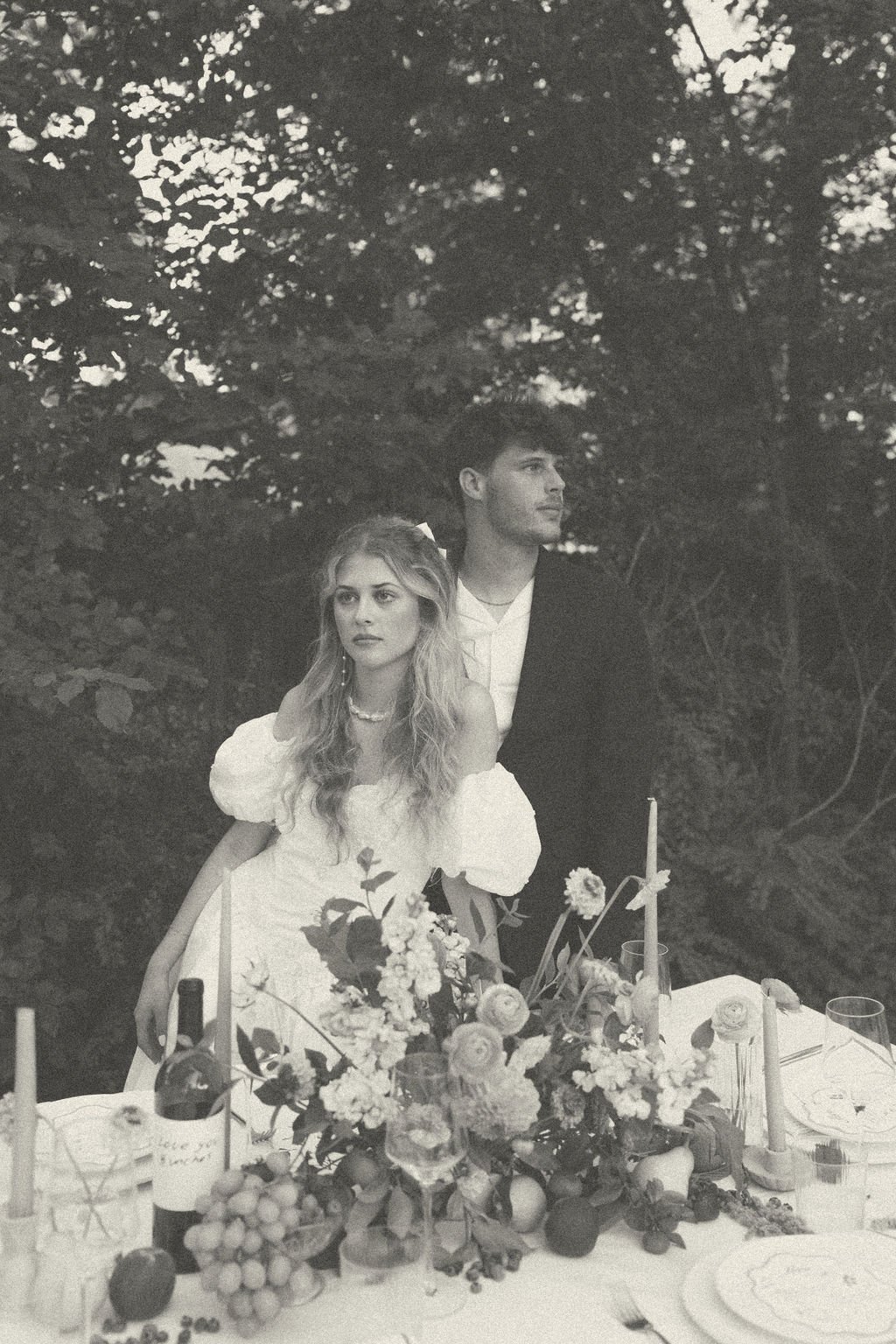 A man and woman standing by an outdoor table decorated with flowers and a bottle of wine, captured in a vintage style black and white photo.