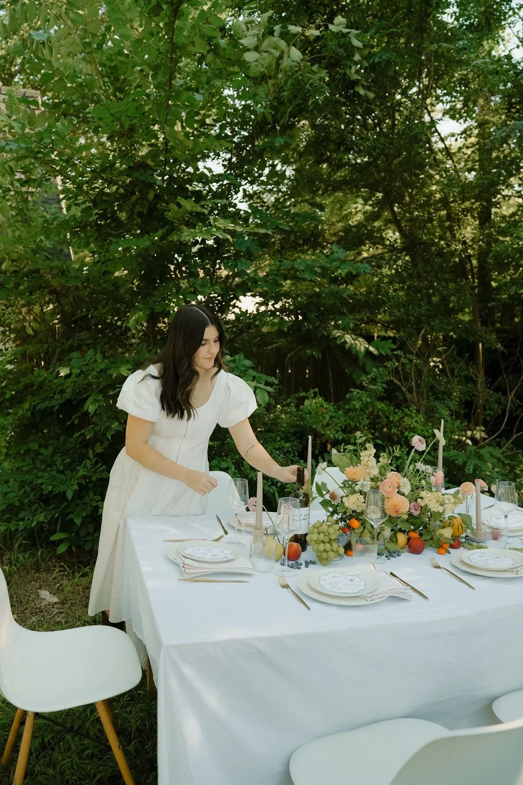 A woman in a white dress setting up a beautifully decorated table outdoors with flowers, fruit, and candles.