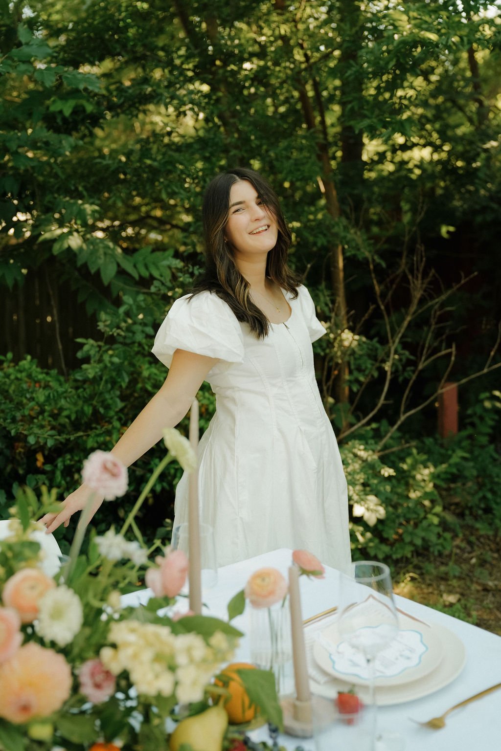 Woman in white dress smiling and standing near a table with flowers and tableware in an outdoor setting.
