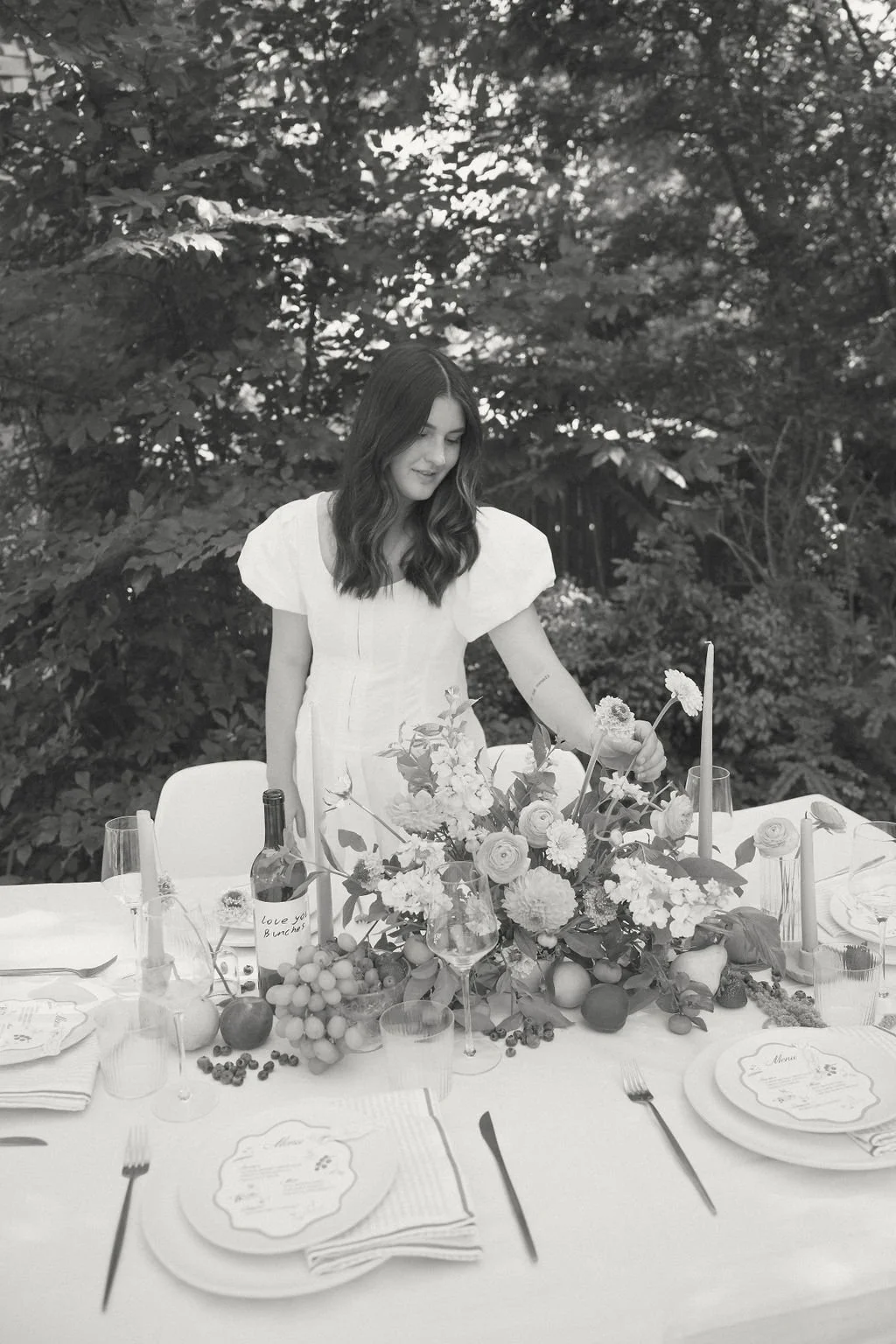 Woman in white dress arranging a floral centerpiece on an elegantly set outdoor dining table with plates, cutlery, and wine glasses.