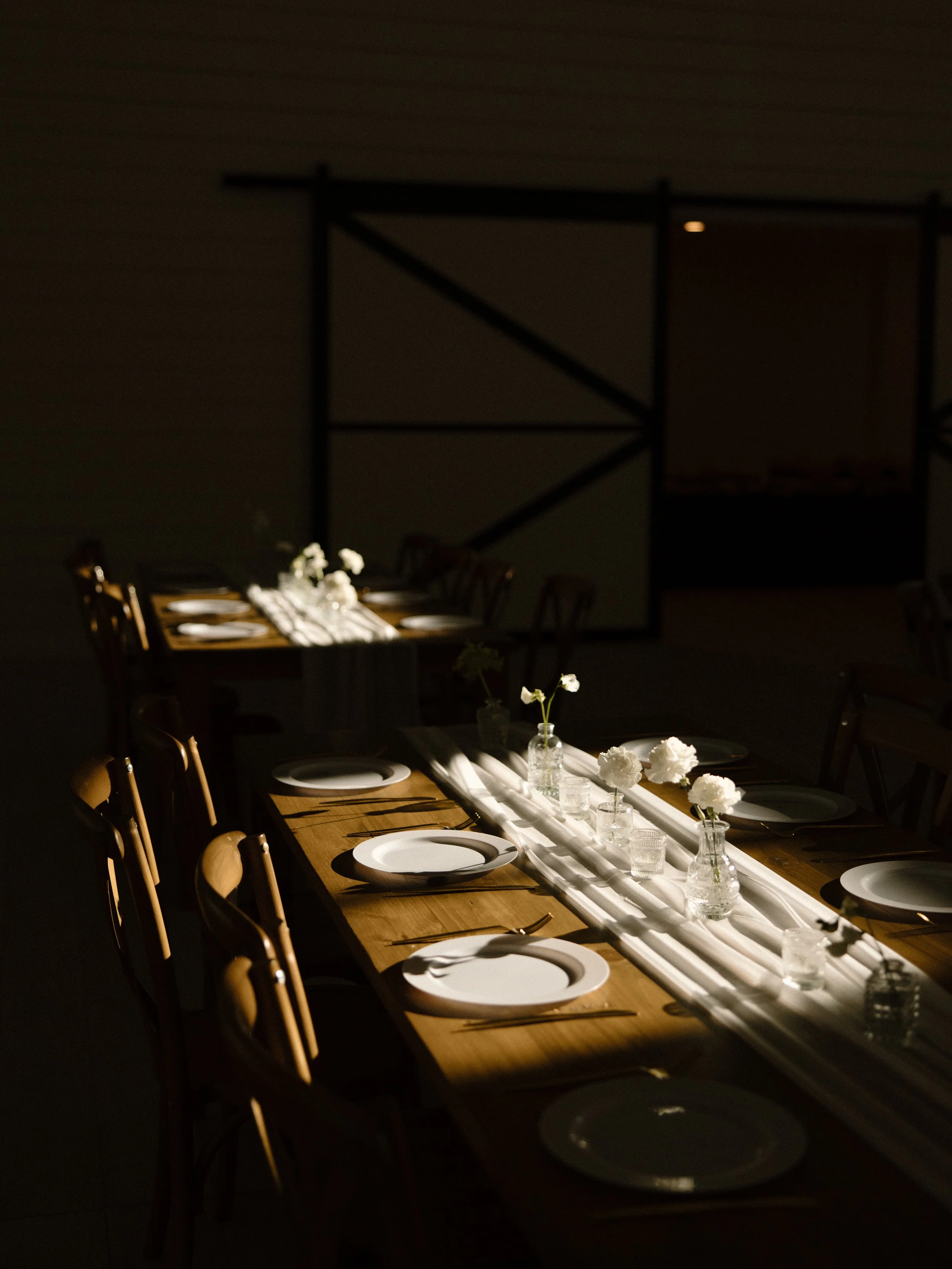 Elegant dining table with white plates, cutlery, and small vases with flowers, in soft lighting.