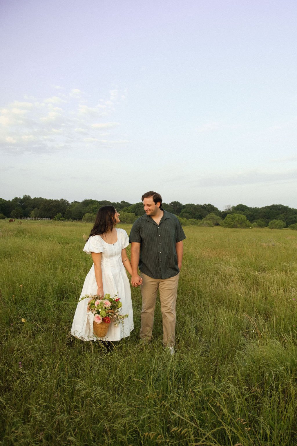 A couple holding hands in a grassy field. The woman is wearing a white dress and holding a bouquet of flowers. The man is in a green shirt and khaki pants. The background includes trees under a clear sky.