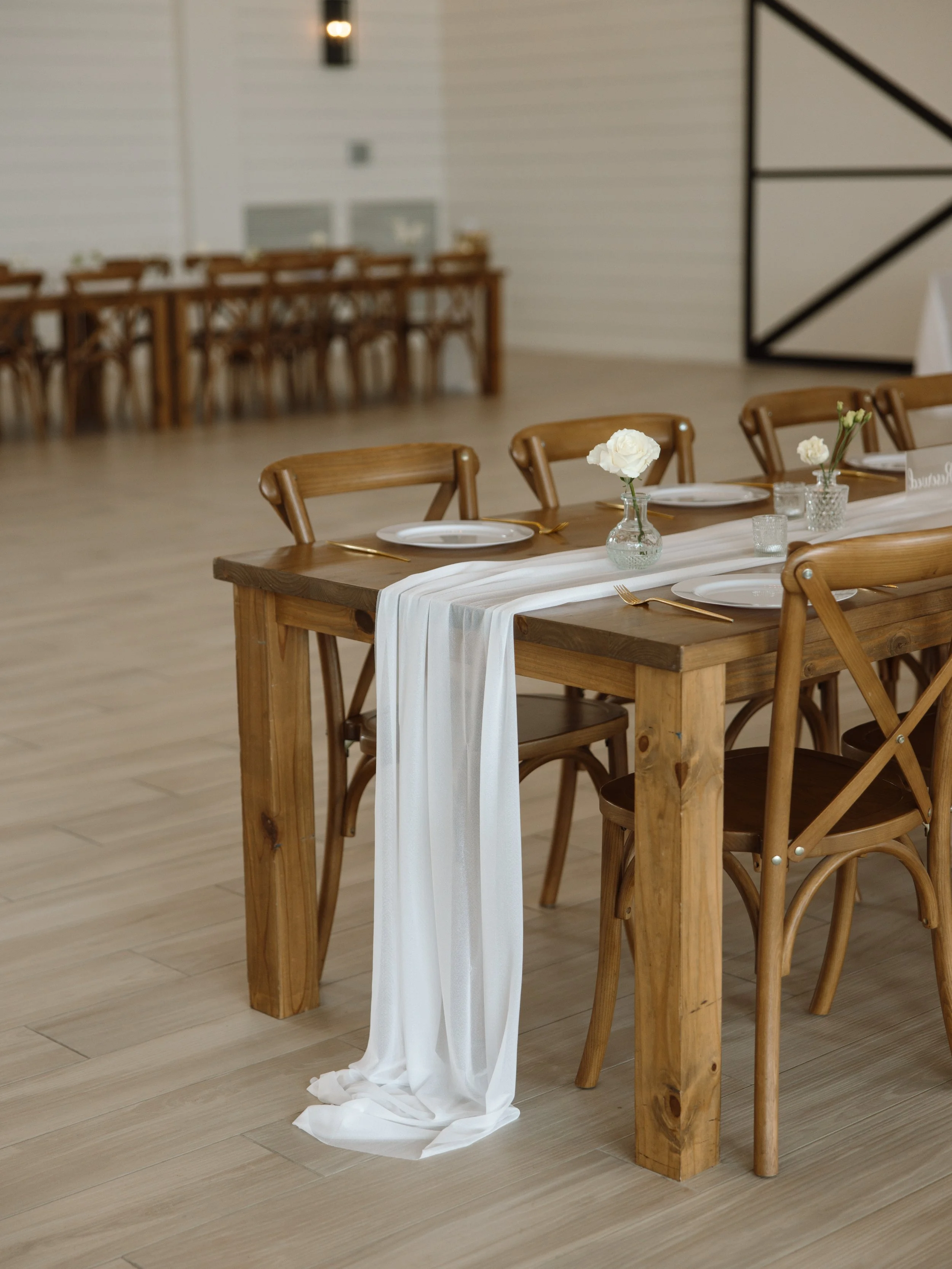 Wedding reception setup with wooden tables, white dinnerware, gold cutlery, and white roses in glass vases.