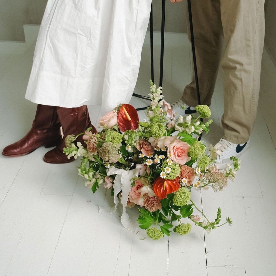 Close-up of two people standing on a white floor, one wearing red boots and a white skirt, and the other wearing beige pants and white sneakers; between them is a bouquet of flowers including roses, anthuriums, and greenery.