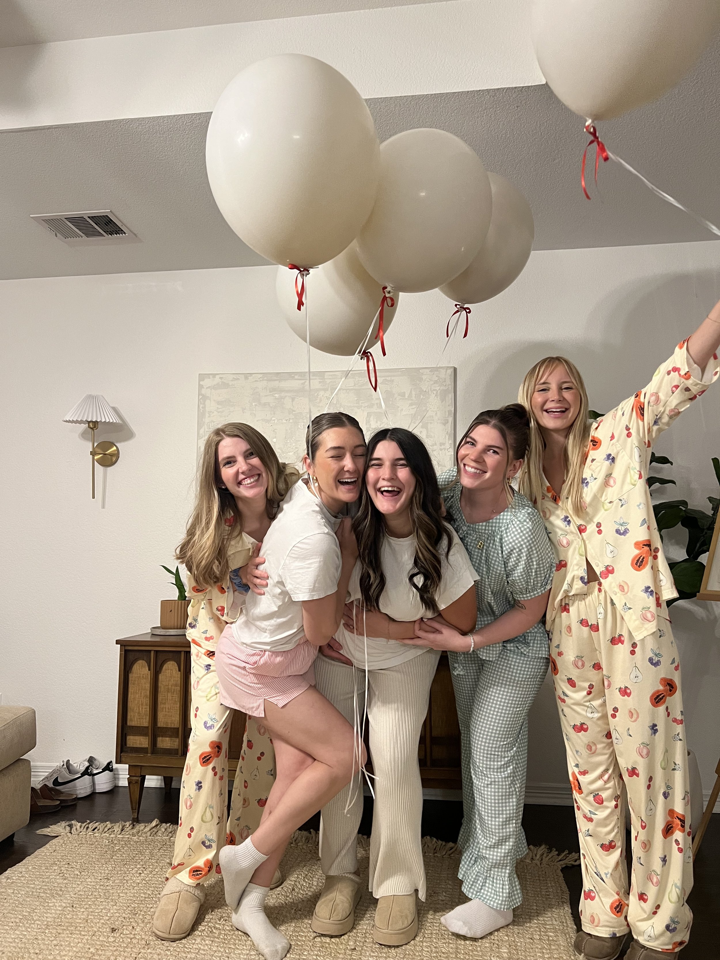 Group of five women wearing pajamas, holding white balloons, smiling in a living room.