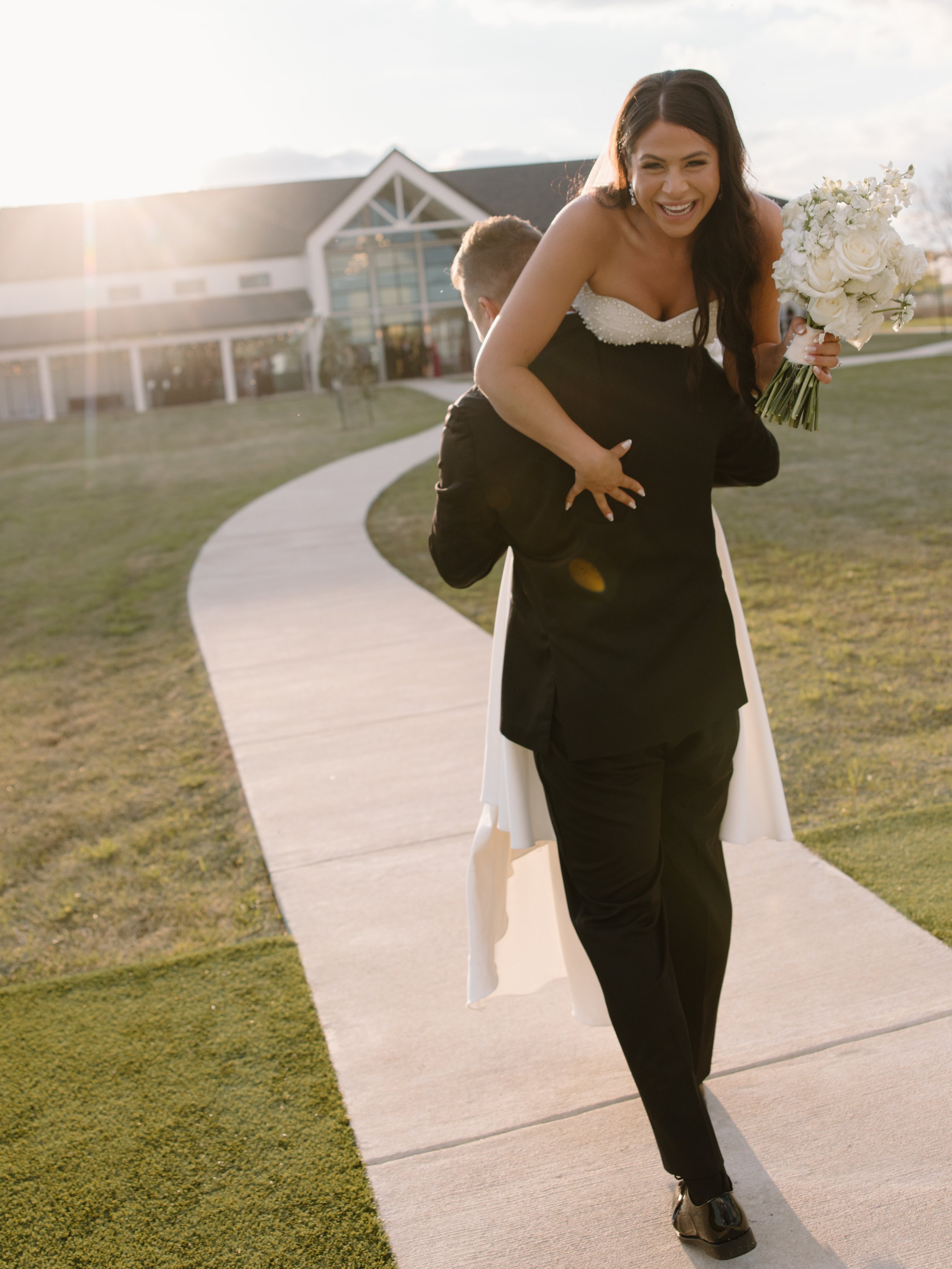 Man carrying woman on his back, woman holding a bouquet, wedding setting, grassy area, building in background.