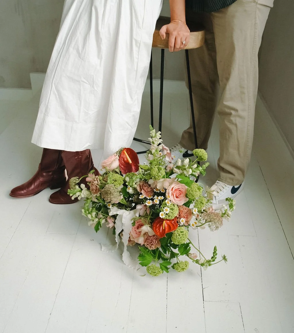 A floral arrangement on the floor with people standing near, one wearing a white dress and brown boots, the other in beige pants and sneakers.