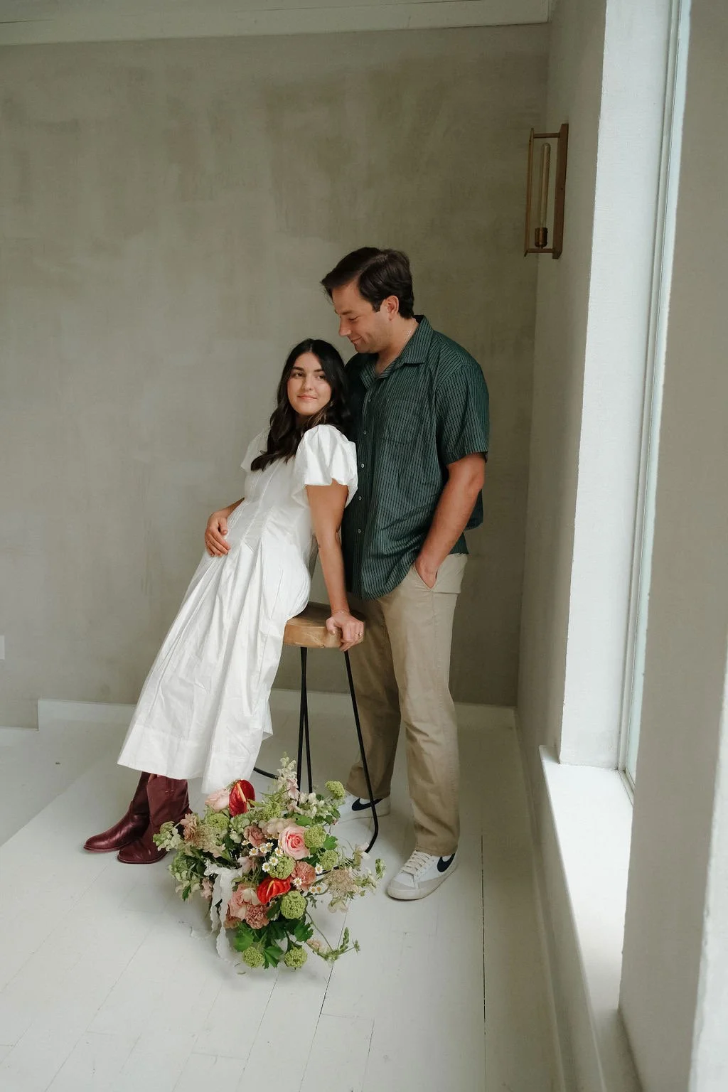 A woman in a white dress sitting on a stool with a man in a green shirt standing beside her. A bouquet of flowers is on the floor near them.