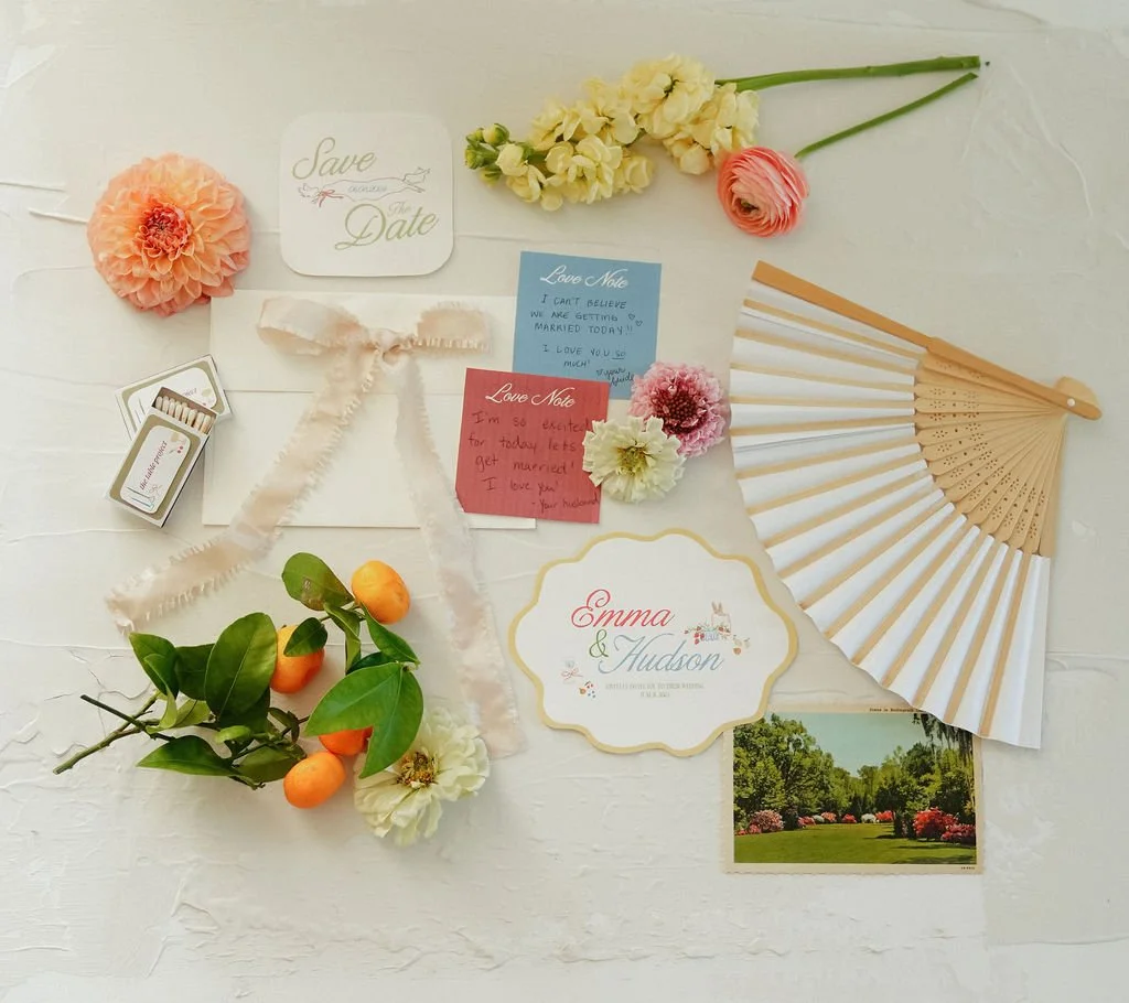 Flat lay of wedding stationery with floral elements, a paper fan, and citrus fruits, including a 'Save the Date' card and love notes for a wedding of Emma and Hudson.
