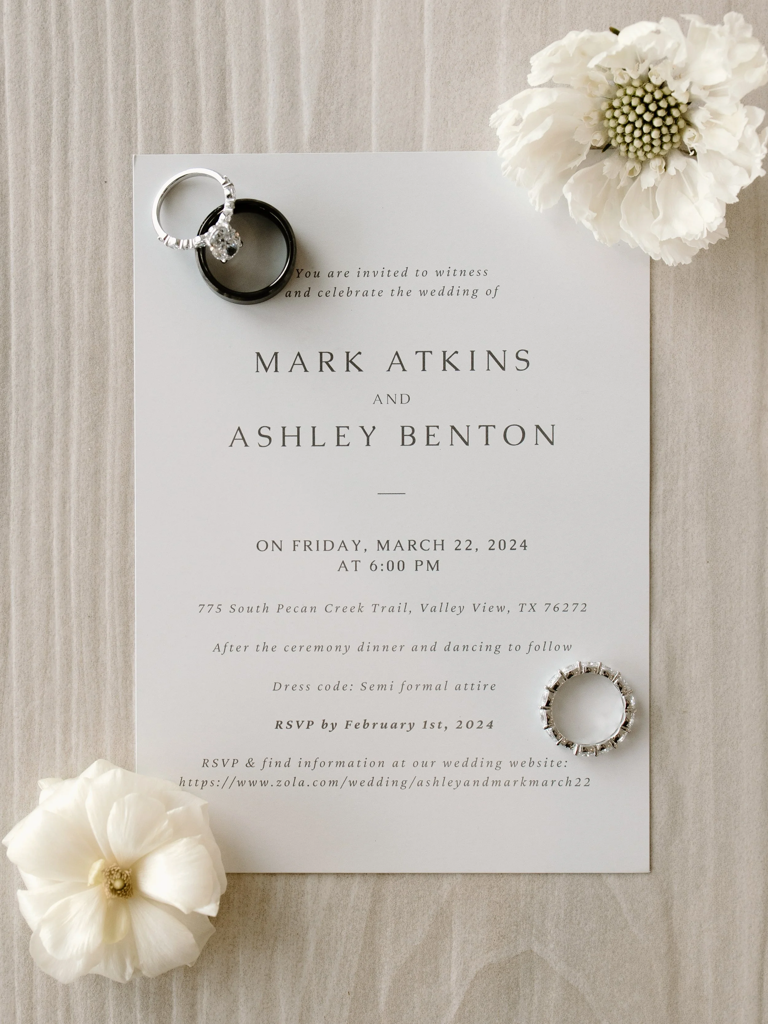 Wedding invitation with rings and white flowers, featuring names Mark Atkins and Ashley Benton, dated March 22, 2024, at 6:00 PM in Valley View, TX.