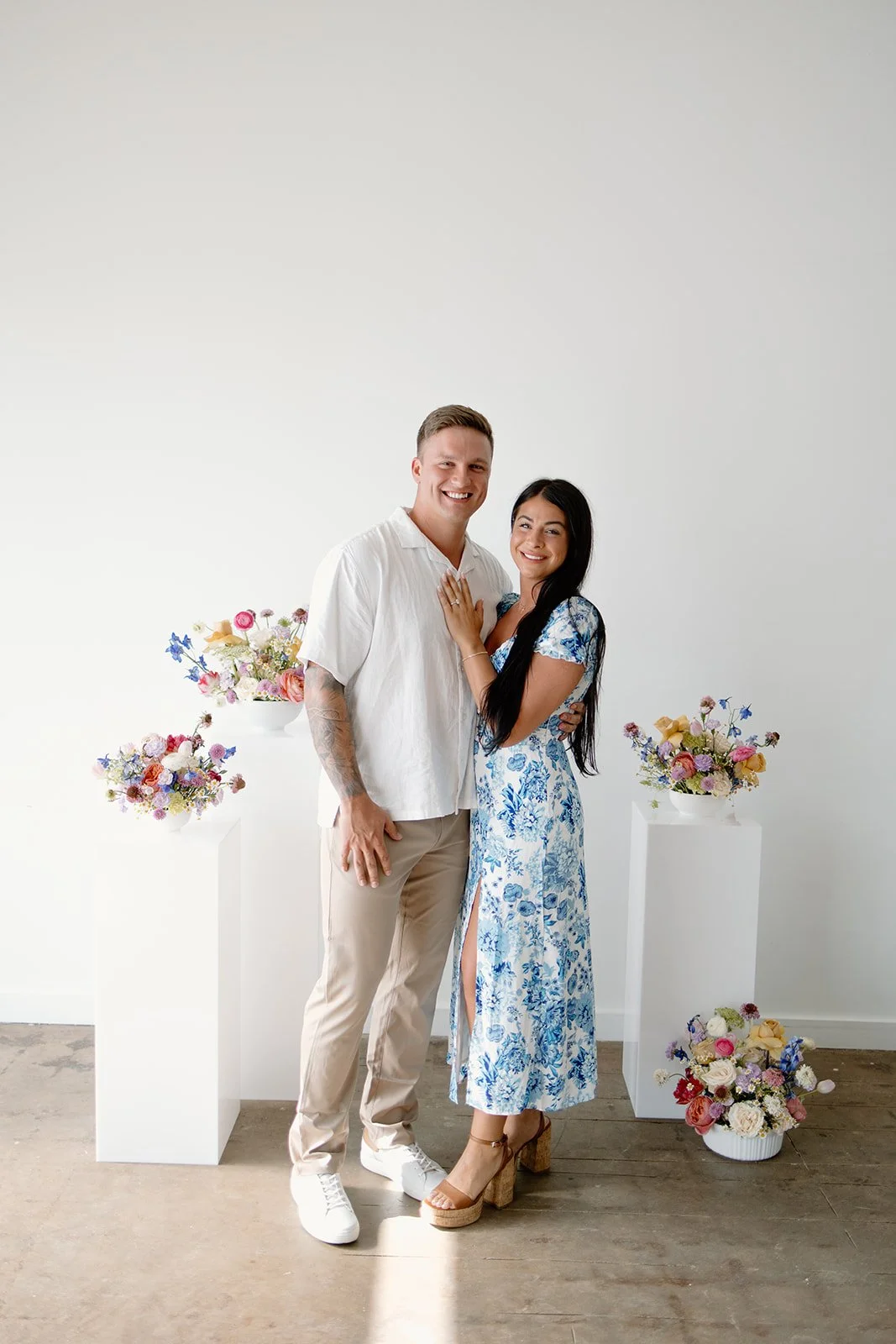 A couple posing together in front of several floral arrangements, with the man wearing a white shirt and beige pants and the woman in a blue floral dress.