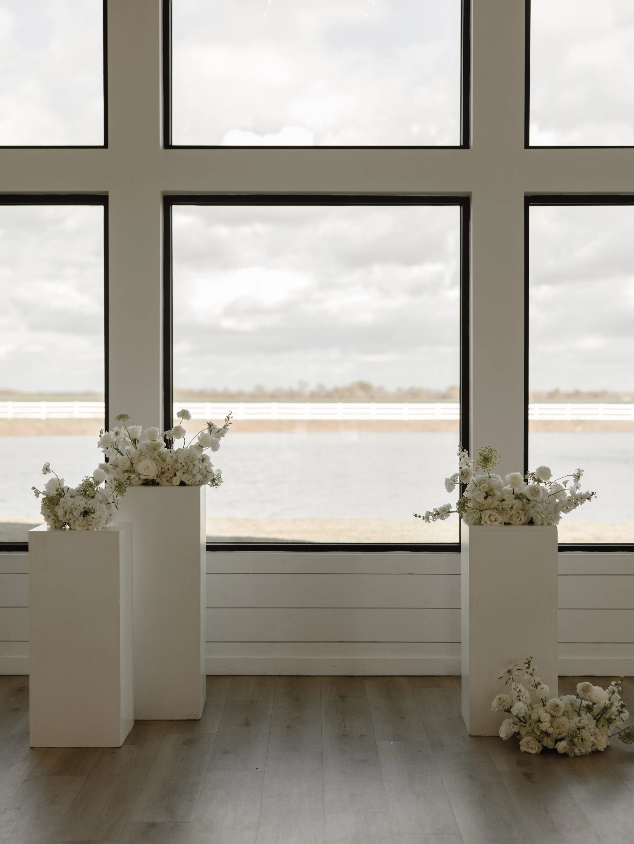 Indoor view of large windows with white floral arrangements on rectangular pedestals, wooden floor, and outdoor landscape visible.