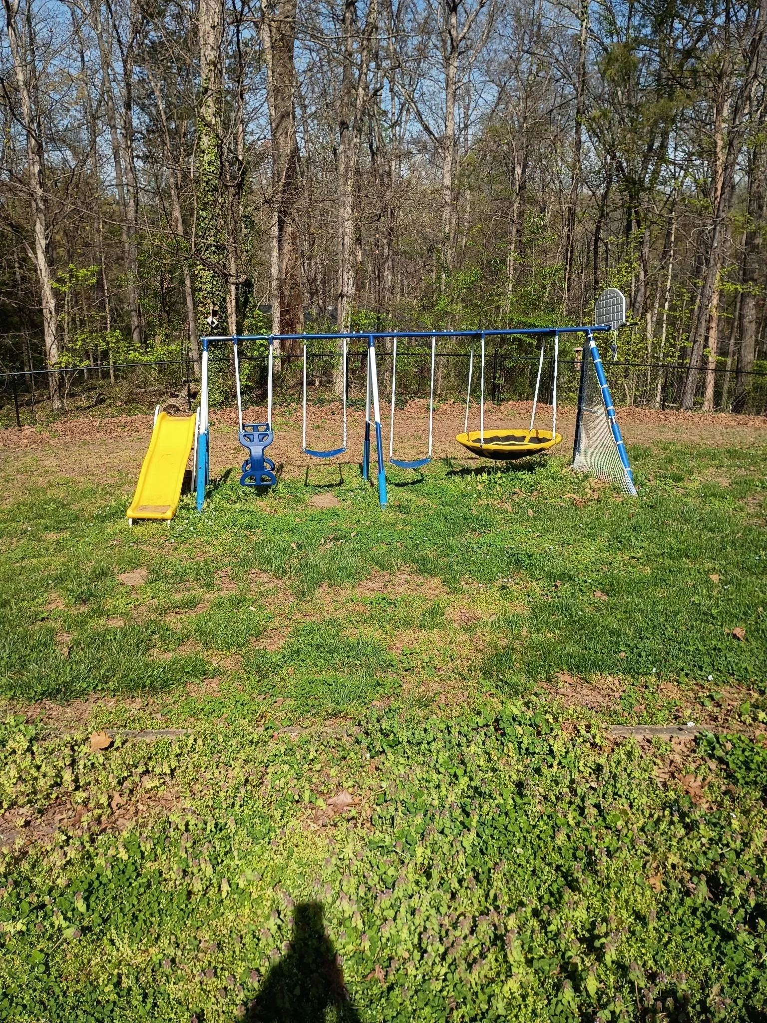 Outdoor playground set with a yellow slide, blue swings, a yellow saucer swing, and a climbing wall, set on a grassy lawn with trees in the background, before clean-up.