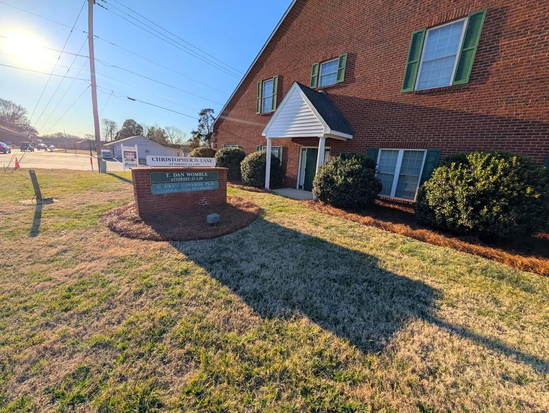 A brick building with green shutters and a small porch, a sign in front lists attorney names, and a grassy lawn with bushes and a shadow cast by the building.